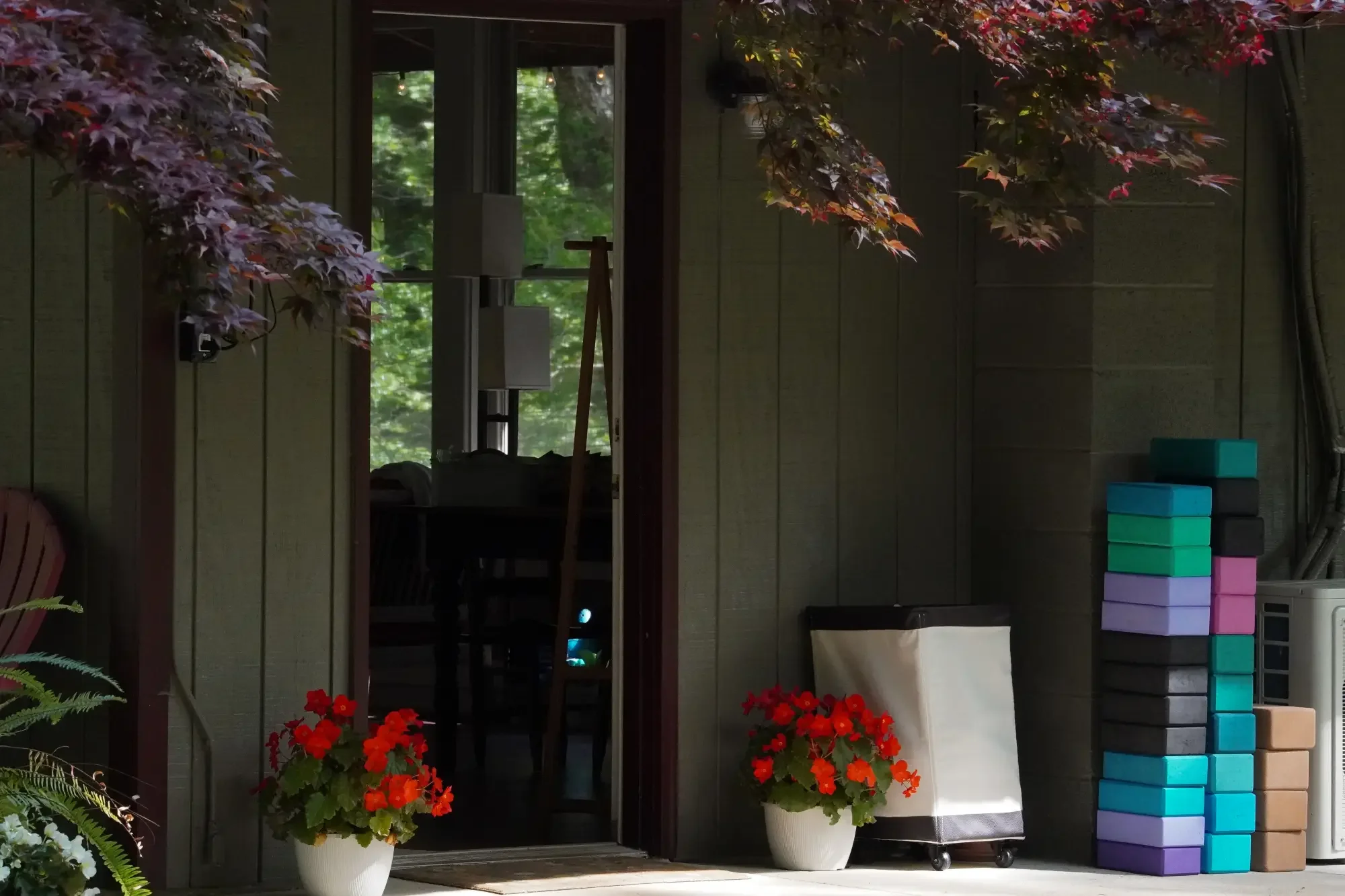 A porch with potted red and orange flowers, colorful therapy or yoga blocks stacked on the right, and a large window revealing green trees outside.
