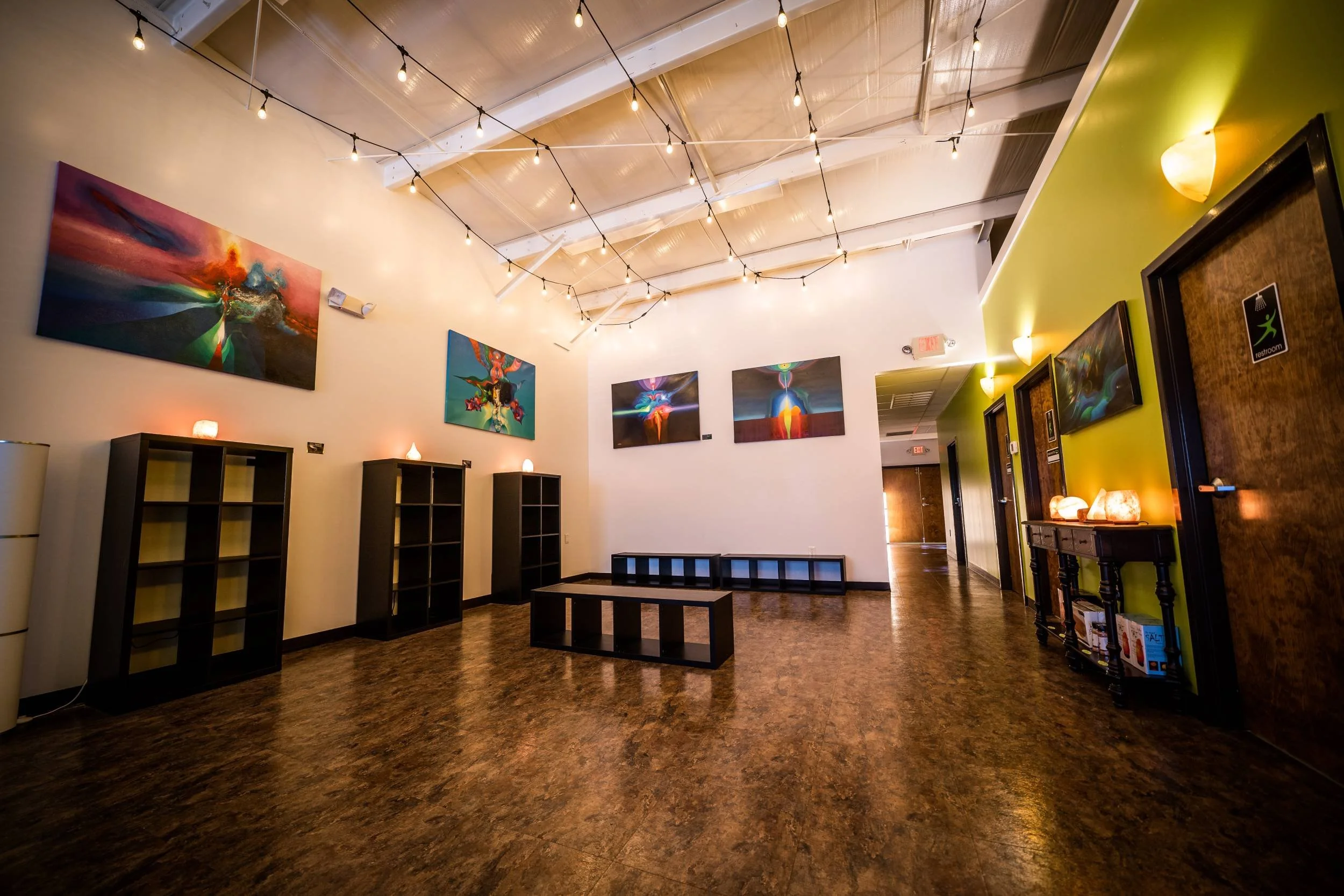 Interior of an art gallery or exhibition space with string lights hanging from the ceiling, colorful abstract paintings on the walls, black shelving units, and warm lighting.