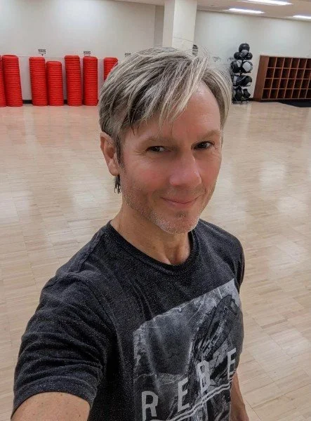 A middle-aged man with light hair and a beard taking a selfie in a gym, with red exercise mats and black fitness equipment in the background.
