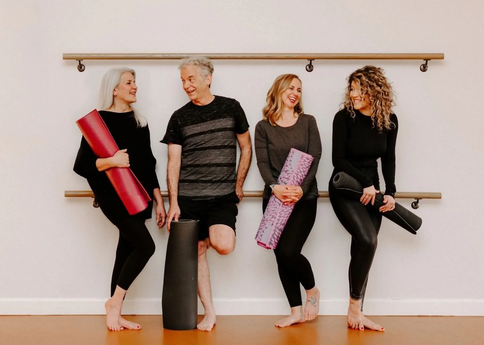 Four women and one man standing against a wall with ballet barres, holding yoga mats and fitness equipment, smiling and engaging with each other, in a fitness or dance studio.