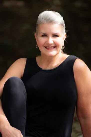 A smiling woman with gray hair styled in an updo, wearing a black sleeveless top, sitting outdoors with a blurred natural background.