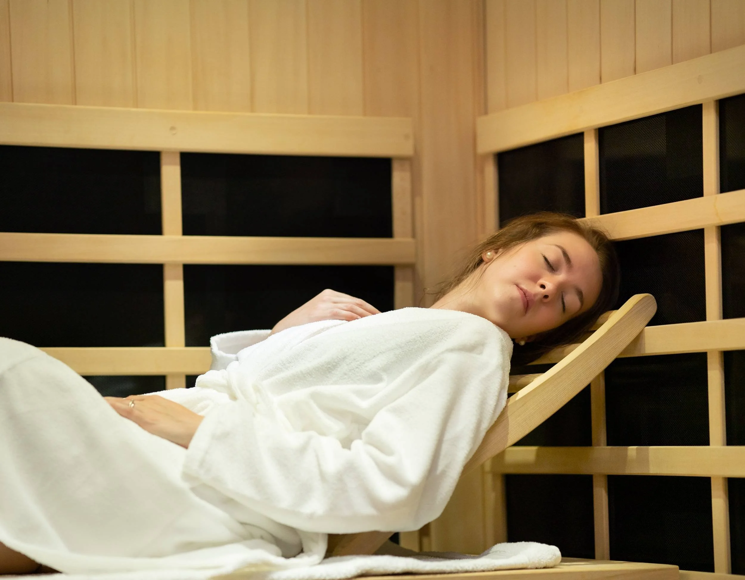A woman with brown hair resting on a wooden recliner with a black acoustic panel behind her, wearing a white towel, in a sauna with wood-paneled walls.