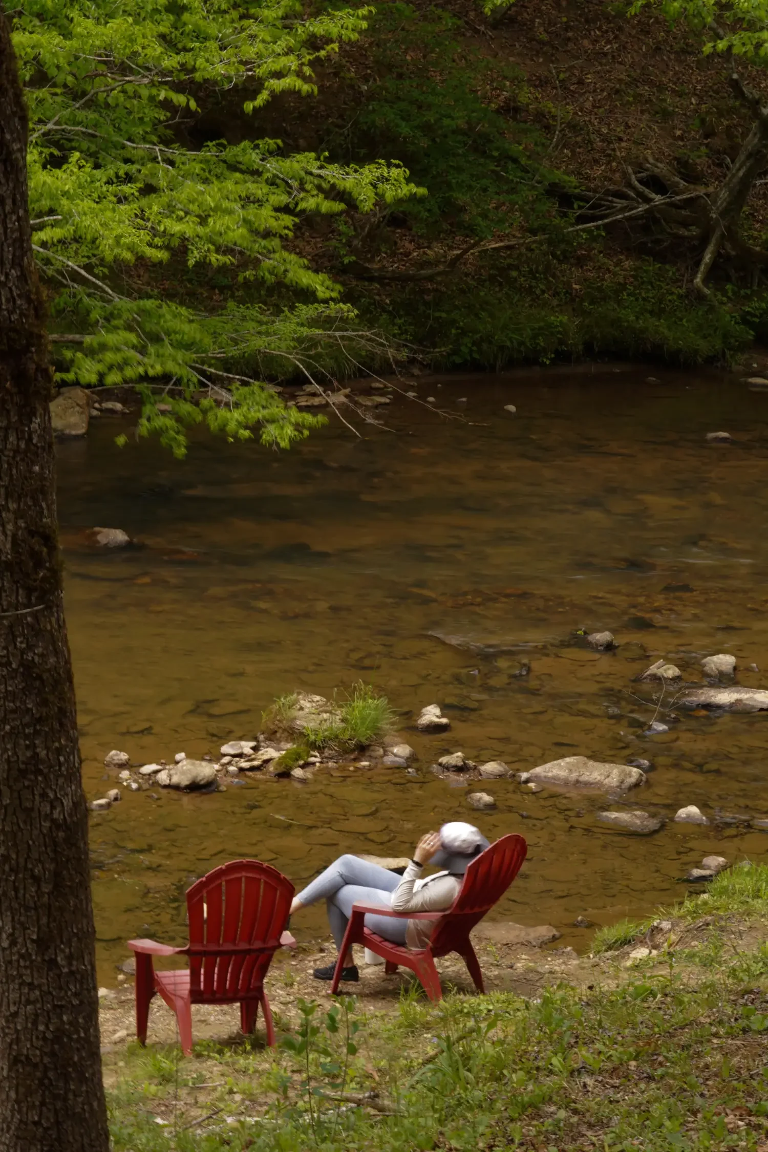 Person relaxing on a red deck chair by a river with trees and rocks in the background.