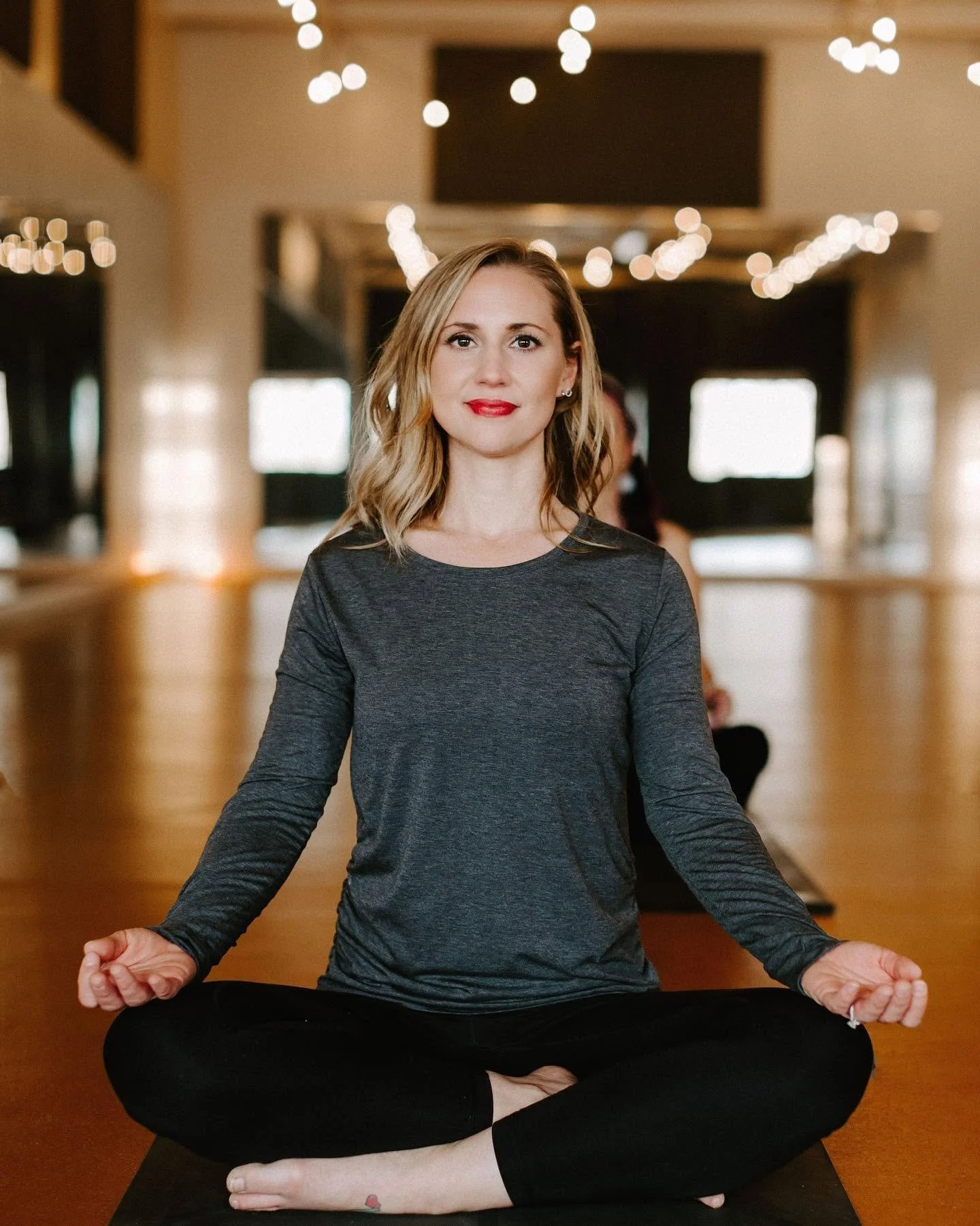 Woman sitting in a cross-legged meditation pose on a yoga mat in a softly lit room with string lights, wearing a dark long-sleeve top and red lipstick.