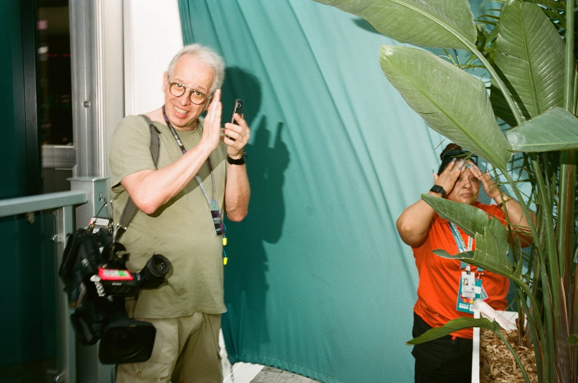 A man with glasses and gray hair, wearing a khaki shirt and holding a camera, is waving to the camera. Next to him, a woman in a red shirt with a badge and lanyard, is standing in front of large green leaves and appears to be covering her face with h