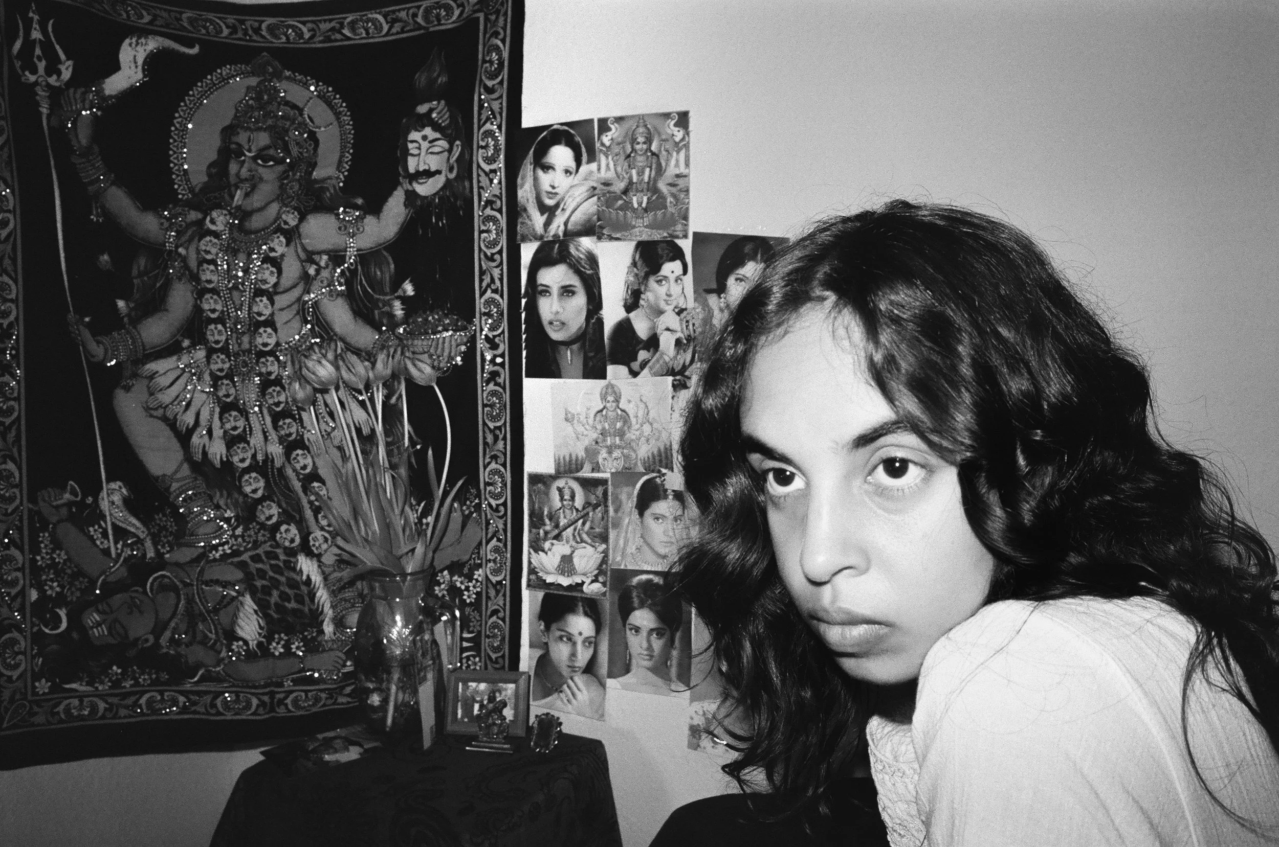 A partial view of a young woman with curly dark hair and light skin, looking at the camera in a room decorated with posters and images of Hindu deities and celebrities on the wall.