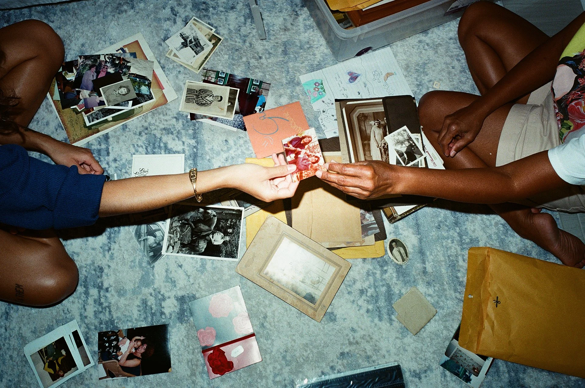 Two people sitting on the floor surrounded by scattered photographs, postcards, drawings, and paper crafts, sharing and exchanging pictures and memories.