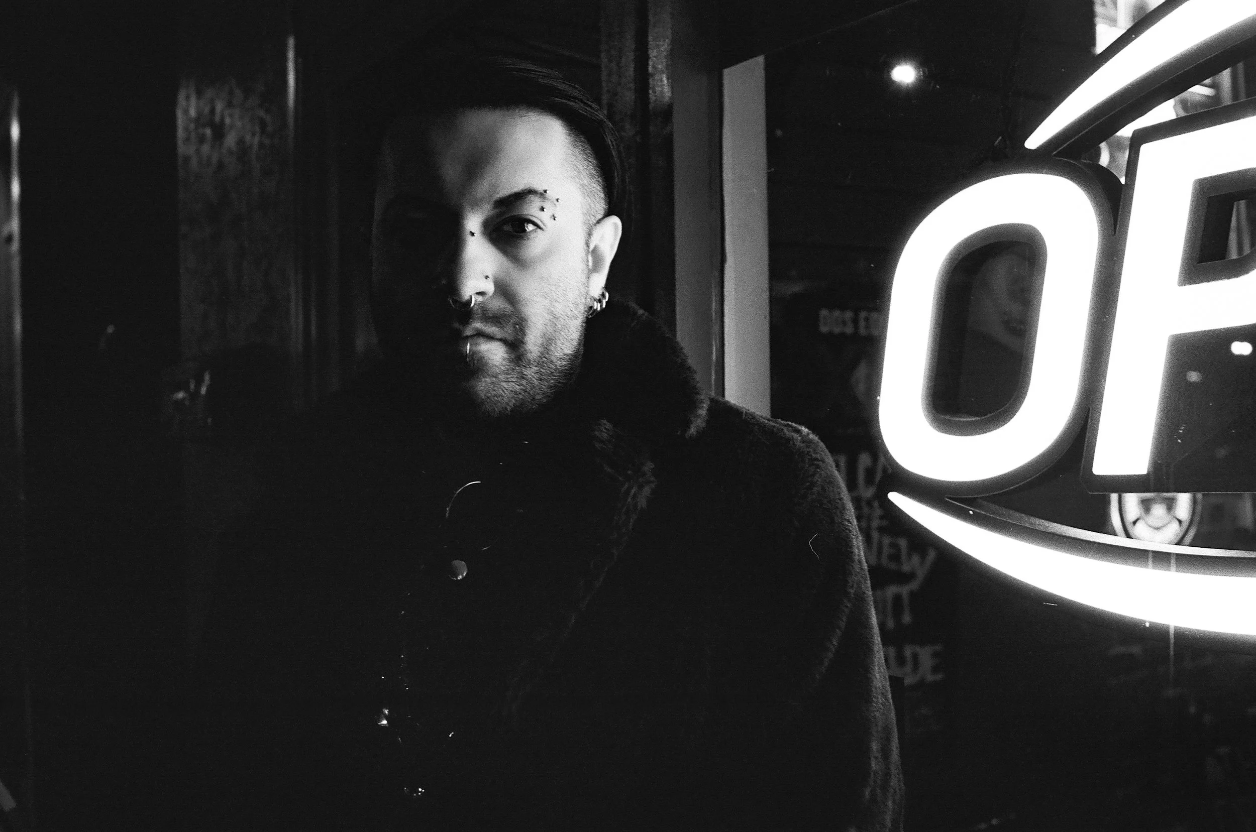 A black-and-white photo of a person with short, dark hair and face piercings looking at the camera, standing near a brightly lit sign with the word 'OPEN' visible in the background.