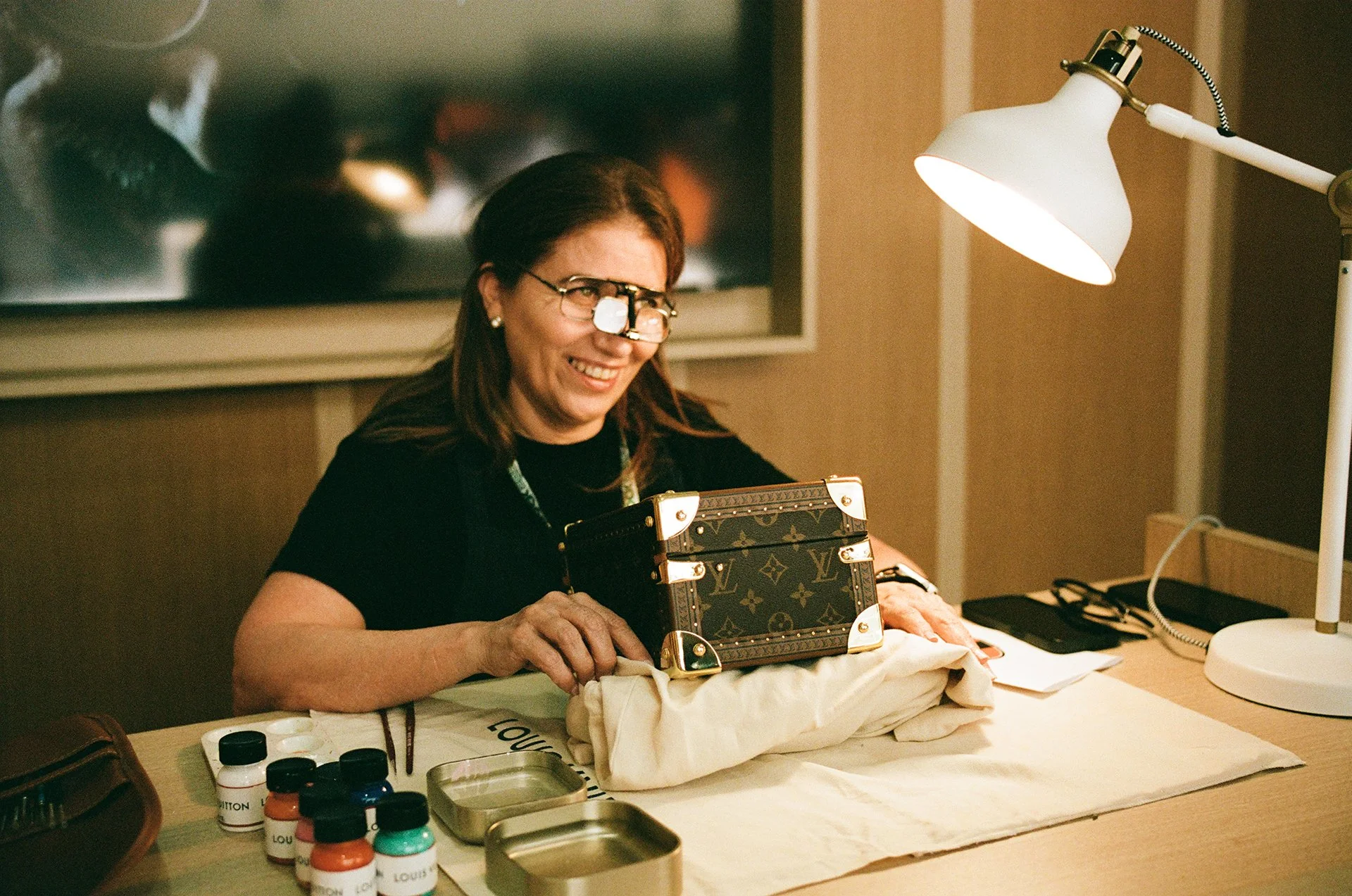 A woman with glasses smiling at a table displays a Louis Vuitton handbag. There are small bottles of paint, paintbrushes, and metal containers on the table. A bright desk lamp illuminates the scene, and a television is visible in the background.