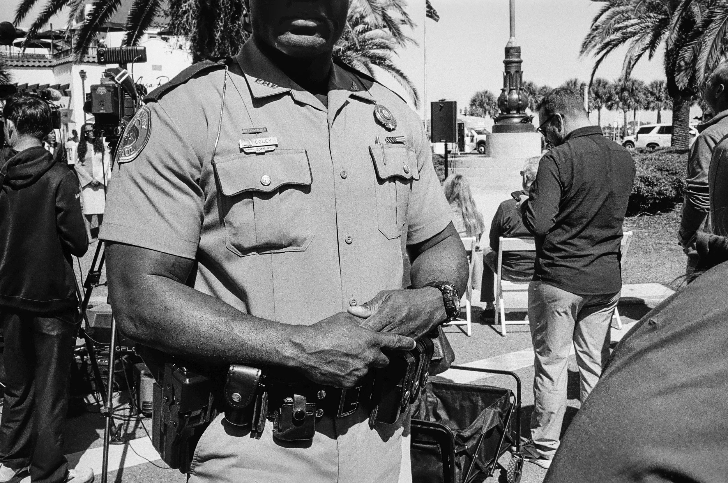 Close-up of a police officer standing outdoors at a public event, with event staff and other individuals in the background, surrounded by palm trees and vehicles.