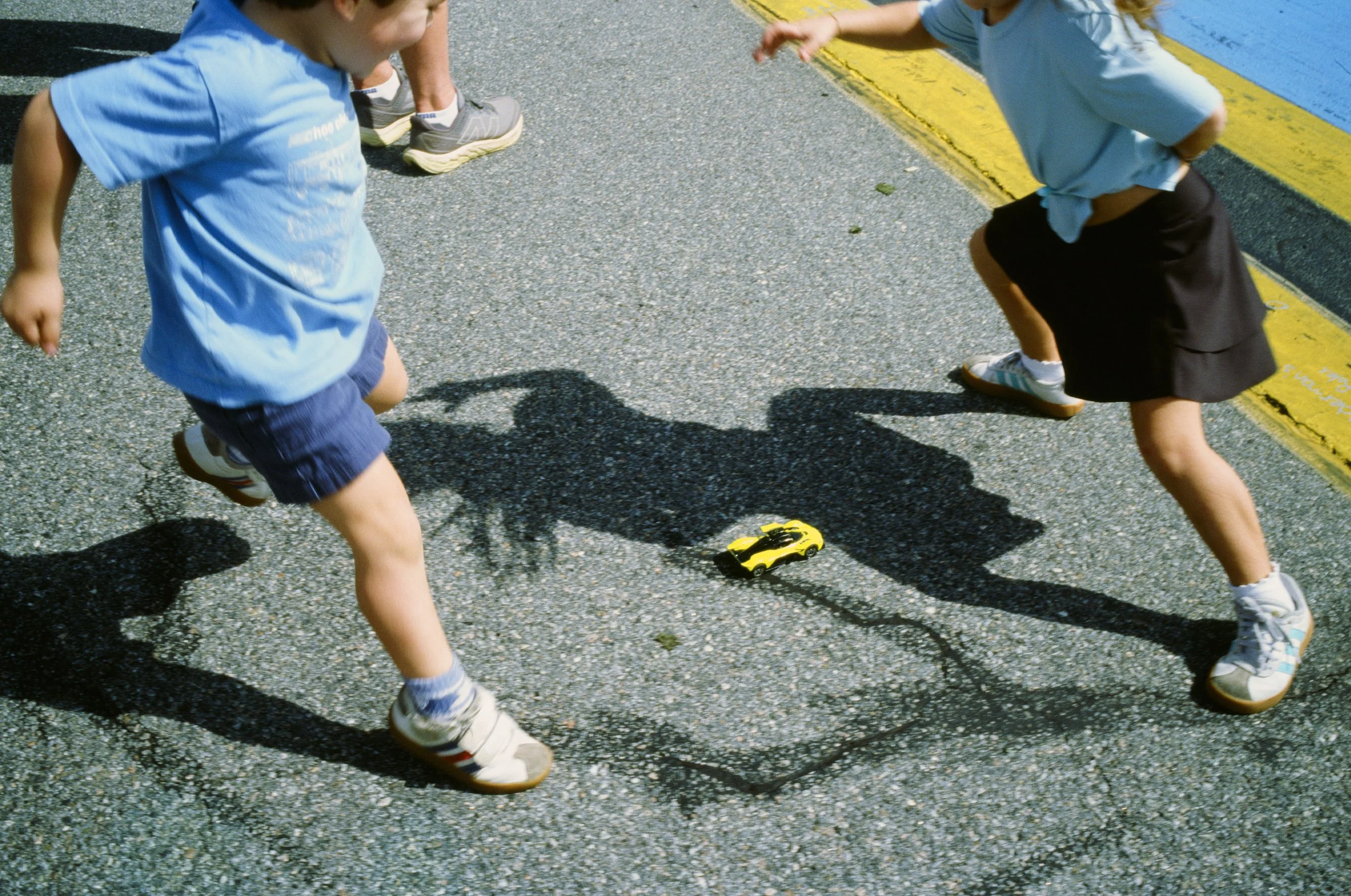 Two children playing with a yellow toy car on the pavement near a painted curb. One child is in a light blue shirt and dark shorts, while the other is in a grey shirt and dark skirt. Shadows are cast on the ground.