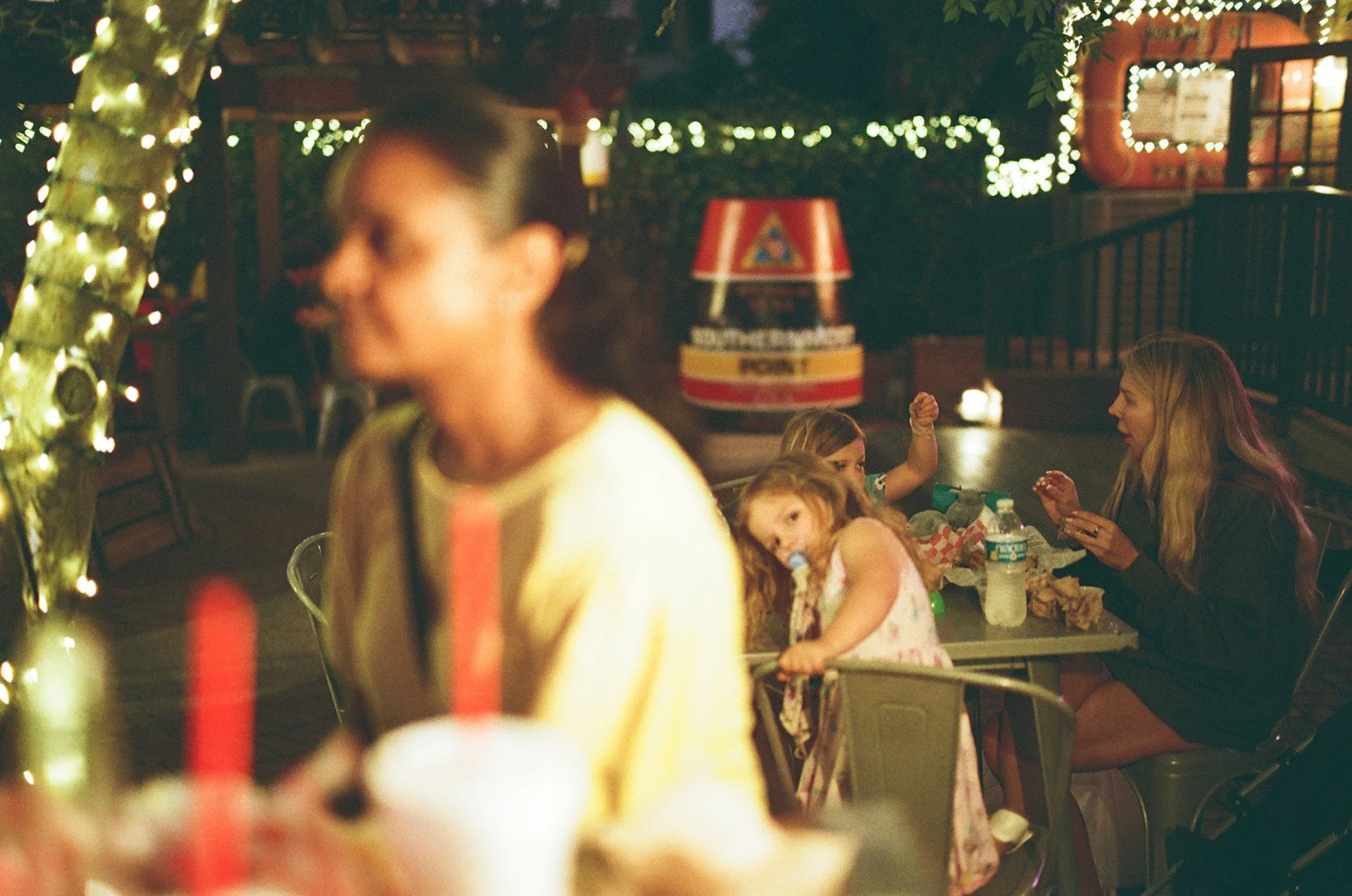 People dining outdoors at night decorated with string lights, with a woman and children at a table, and a blurred woman in the foreground.