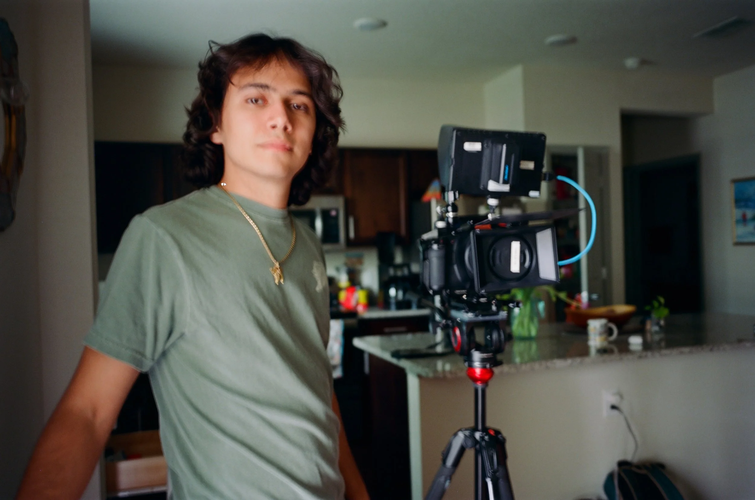 A young man with shoulder-length dark hair wearing a green T-shirt and gold chain stands next to a camera mounted on a tripod inside a home.