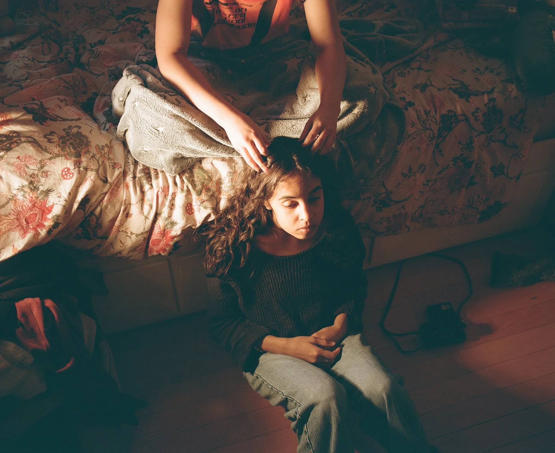 A person with curly hair lying on a bed with floral sheets, eyes closed, resting or sleeping, while another person appears to be petting or adjusting their hair.