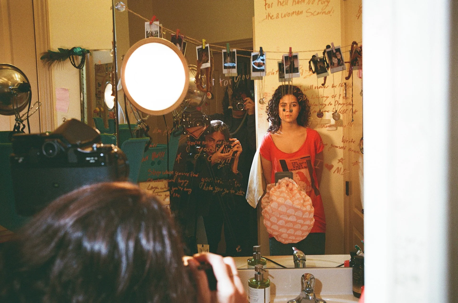 Young woman with curly hair taking a selfie in front of a mirror, holding a pink patterned bag, wearing a red T-shirt, in a room decorated with photos and writing on the wall.