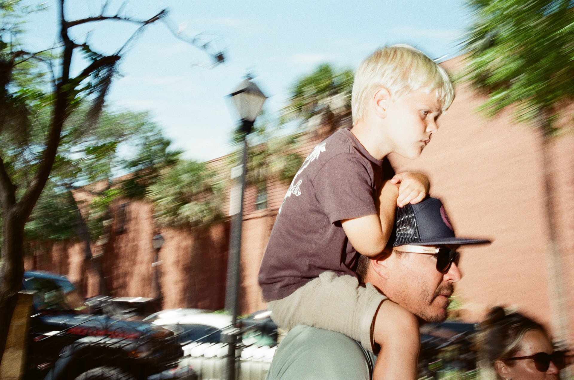 A young boy with blonde hair sitting on one man’s shoulders, holding onto his head, while another man wearing sunglasses supports him from below. They are outdoors with trees, a brick wall, and parked cars in the background.