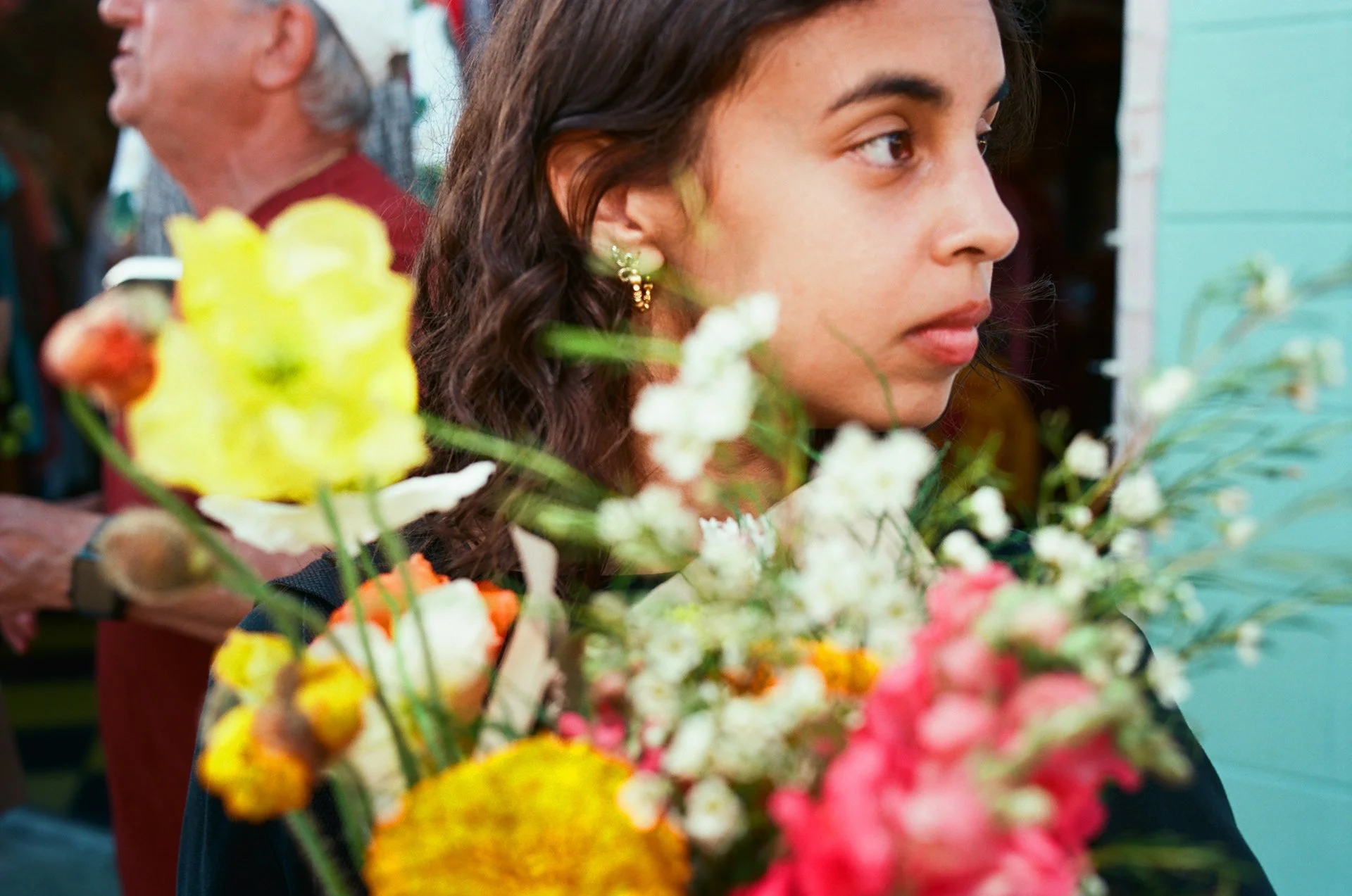 A woman with dark hair and earrings is near a colorful flower arrangement, with a man in a red shirt in the background.