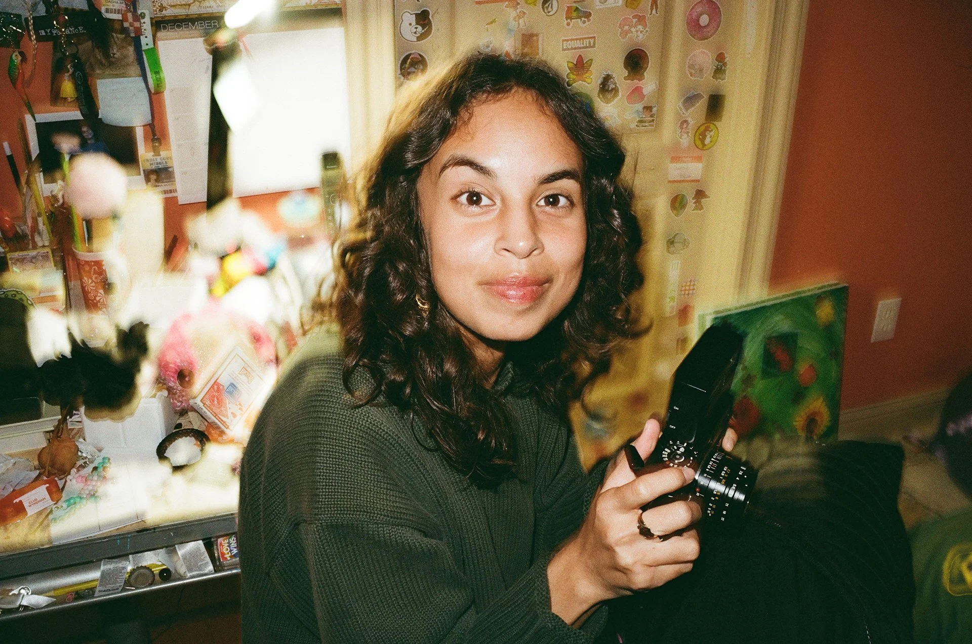 A woman with curly dark hair holding a vintage camera inside a room decorated with colorful artwork and stickers on the wall, with a counter displaying various small items.