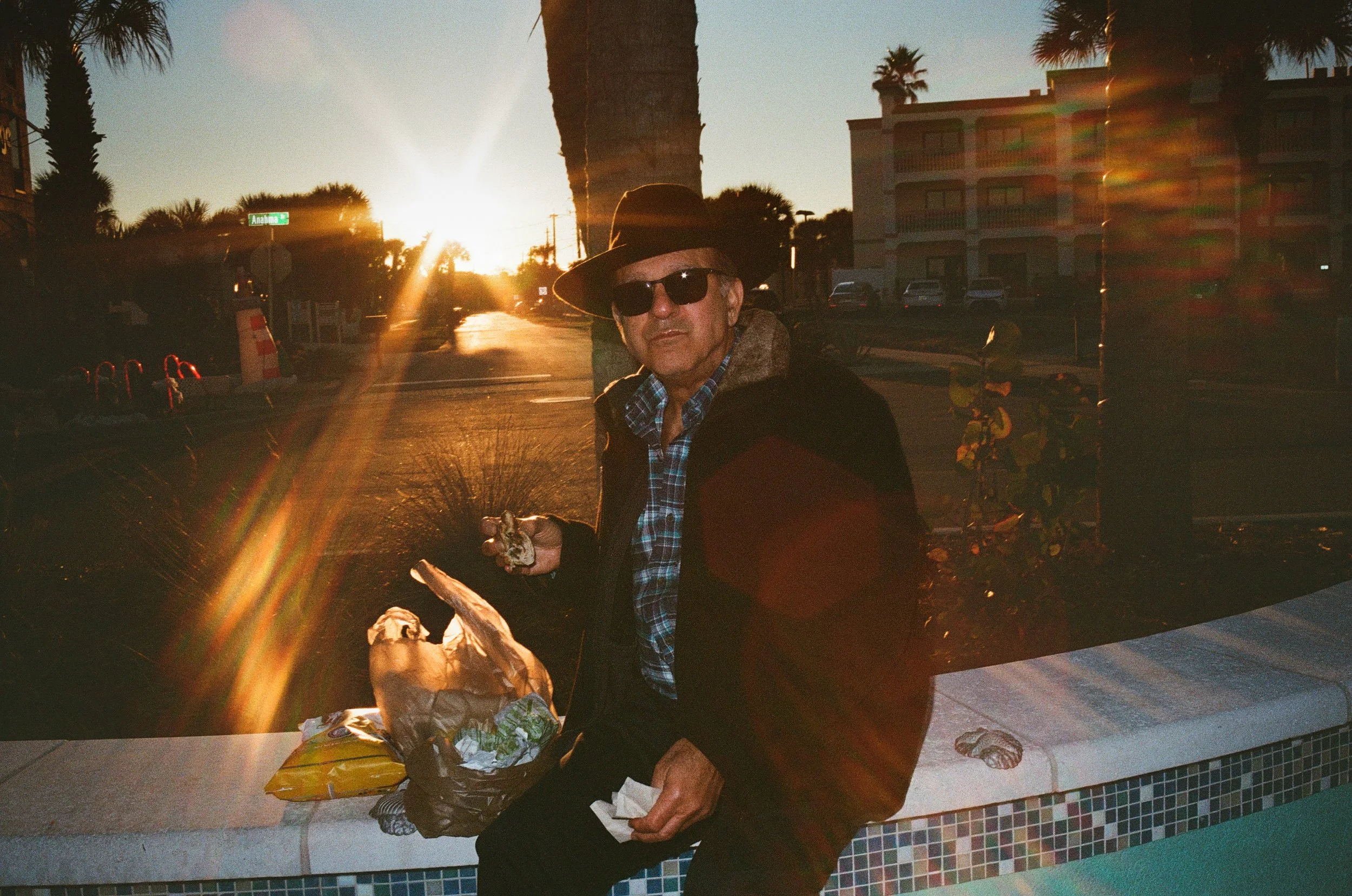 A man wearing a wide-brimmed hat and sunglasses sitting at a table outdoors during sunset, eating food with a paper and plastic bag on the table. Palm trees and buildings are visible in the background.