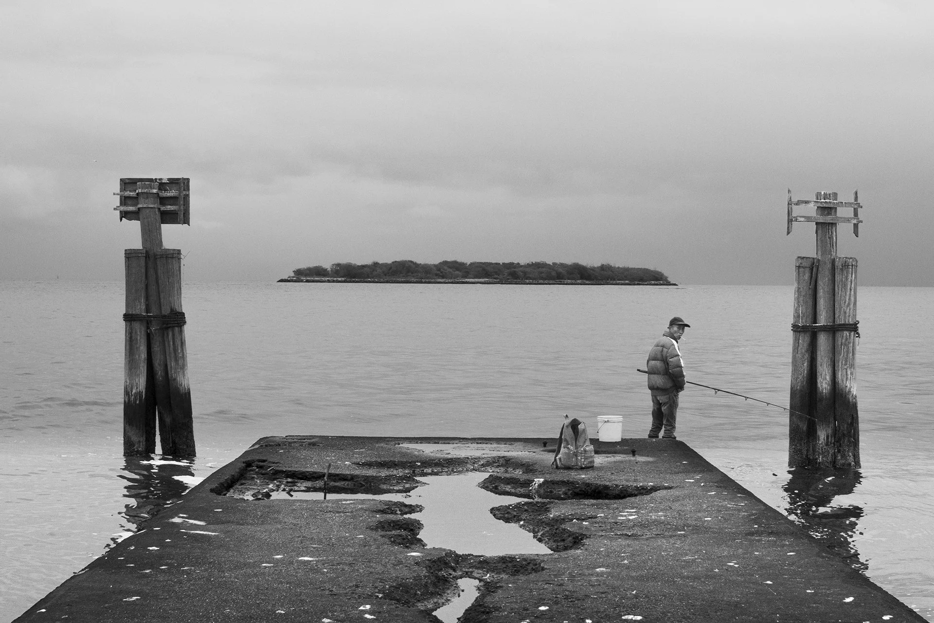 A man fishing on a pier next to two wooden posts on water, with small islands in the background on an overcast day.