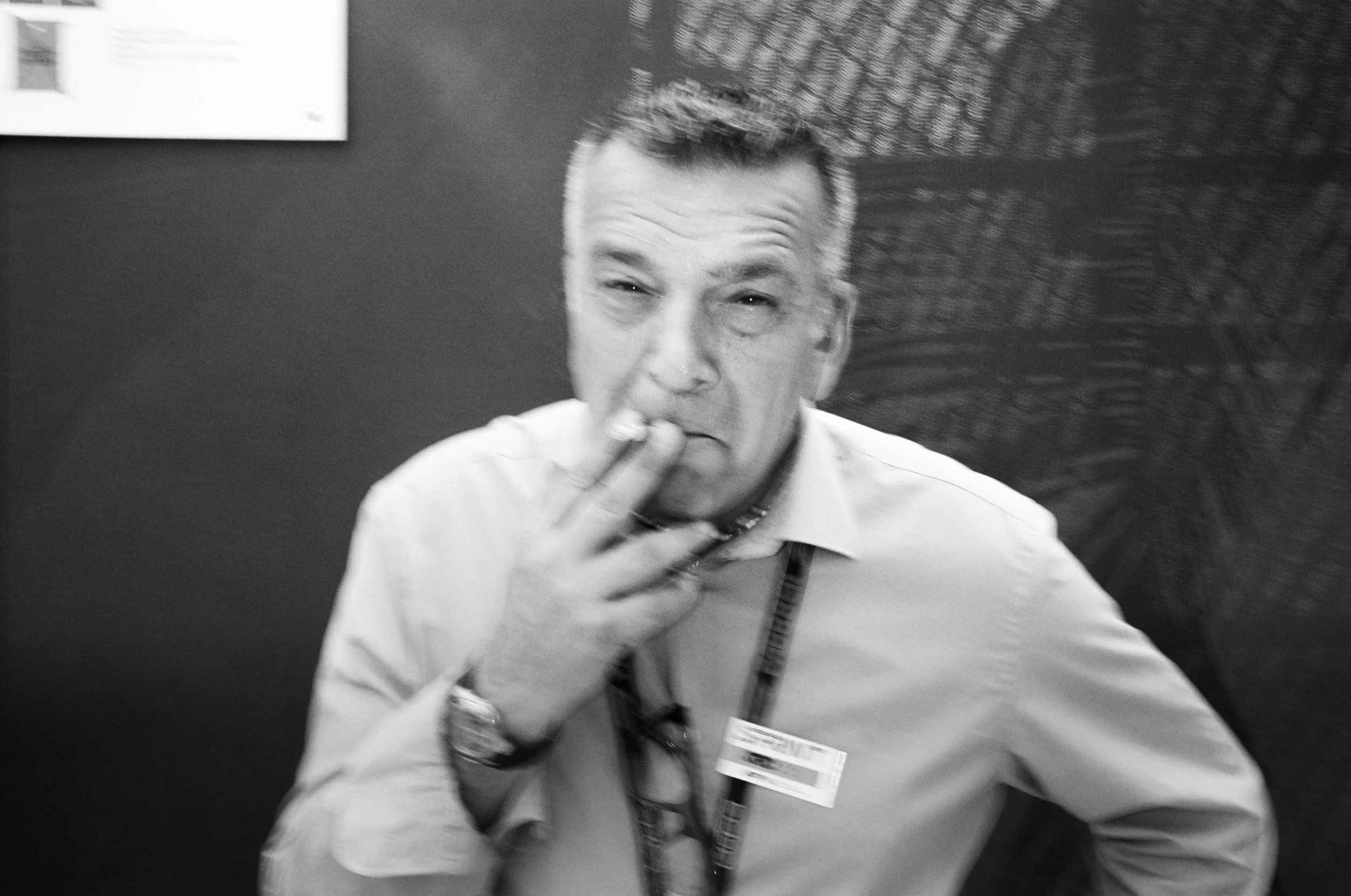A black and white photo of a man with short hair smoking a cigarette. The man is wearing glasses and a collared shirt with a lanyard and ID badge. The background has a textured wall and a framed picture.