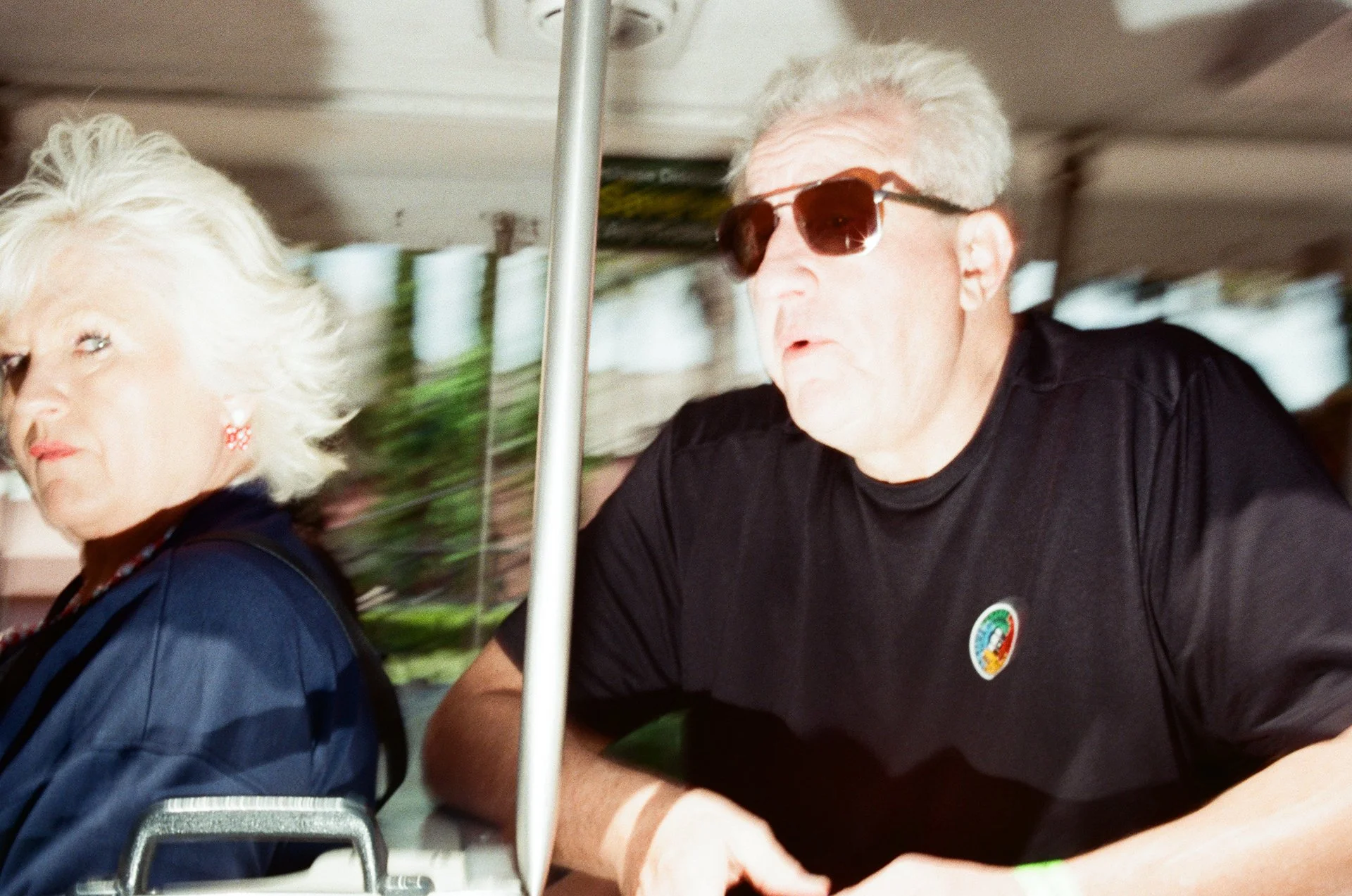 A man and an older woman sitting together on a bus or tram. The man is wearing sunglasses and a black t-shirt with a colorful logo. The woman has white hair, earrings, and is wearing a dark blue or black outfit. They appear to be engaged in an activi