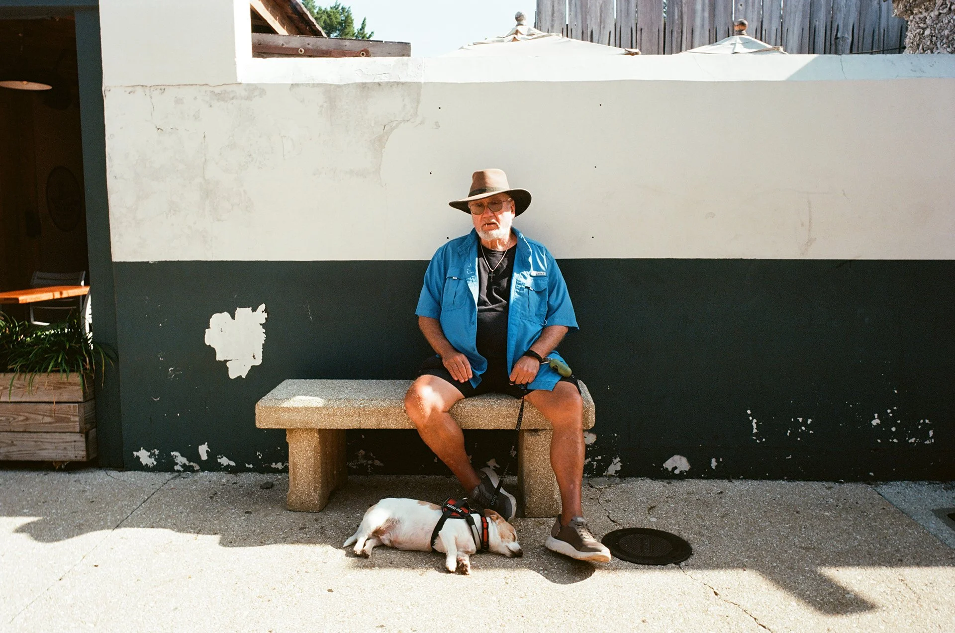 An elderly man with a white beard, wearing a wide-brimmed hat, sunglasses, a blue short-sleeve shirt, and shorts, is sitting on a stone bench with his left leg crossed over his right knee. He has a leash attached to a sleeping dog lying on the ground