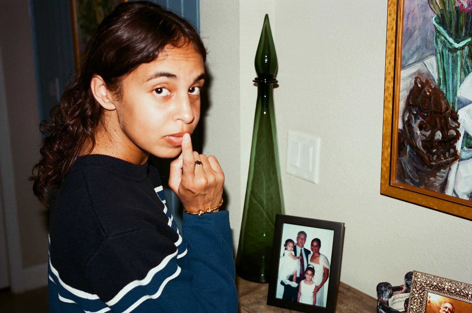 A woman with dark brown curly hair, wearing a dark blue sweater with white and blue stripes, is touching her lips with her index finger, beside a framed family photo, a tall green glass decorative bottle, and a painting of flowers in a vase on the wa