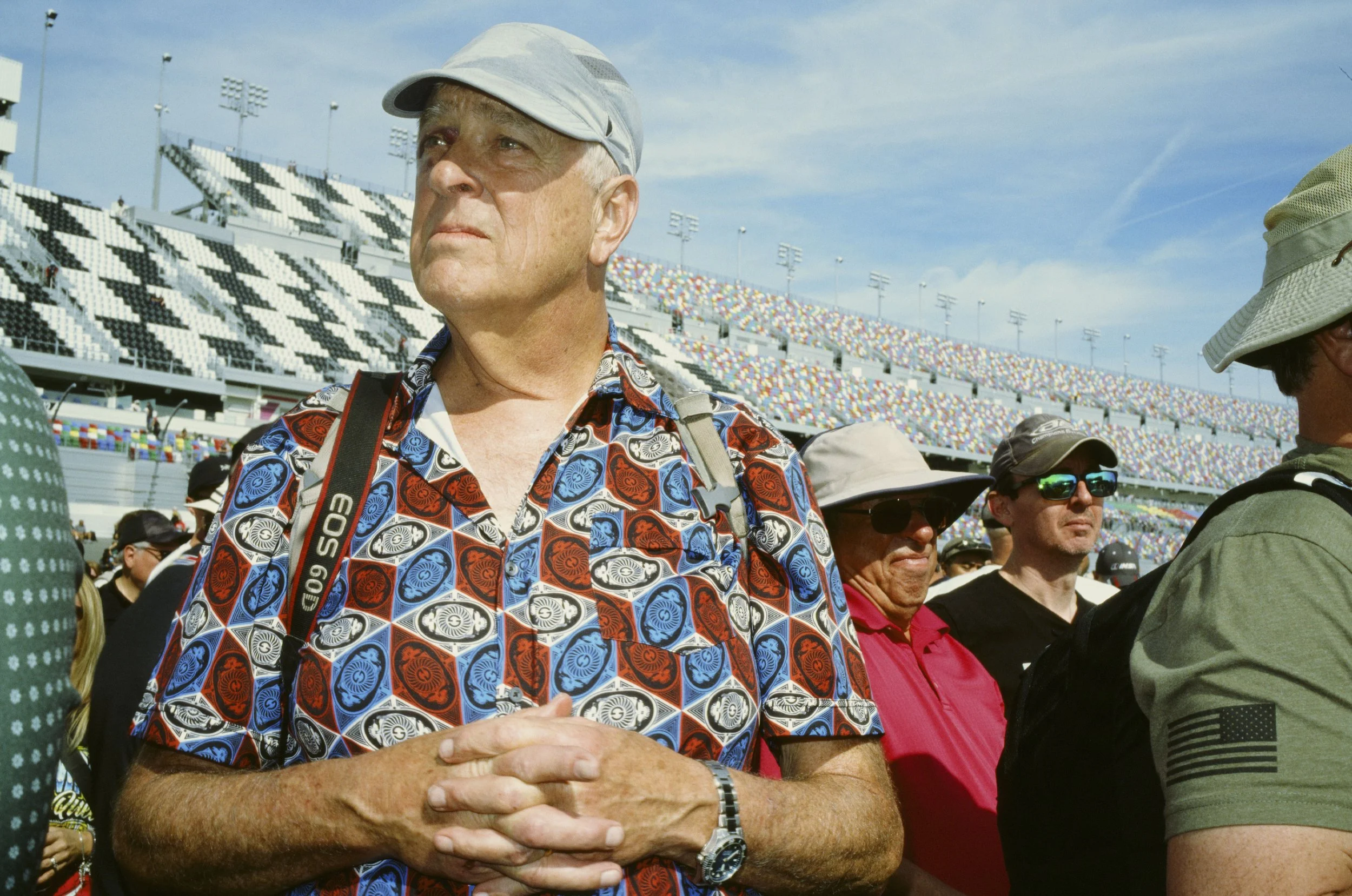 A man in a colorful patterned shirt and cap standing among a crowd at a sports stadium.