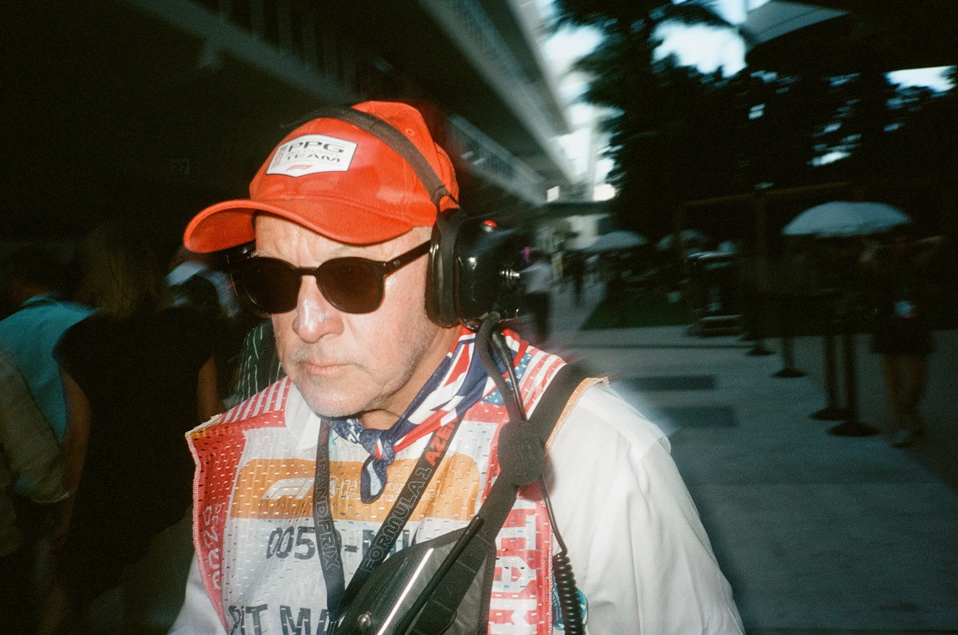 A man wearing a red cap, sunglasses, and a racing vest appears to be in a busy outdoor setting, possibly an event or gathering, with trees and a building in the background.