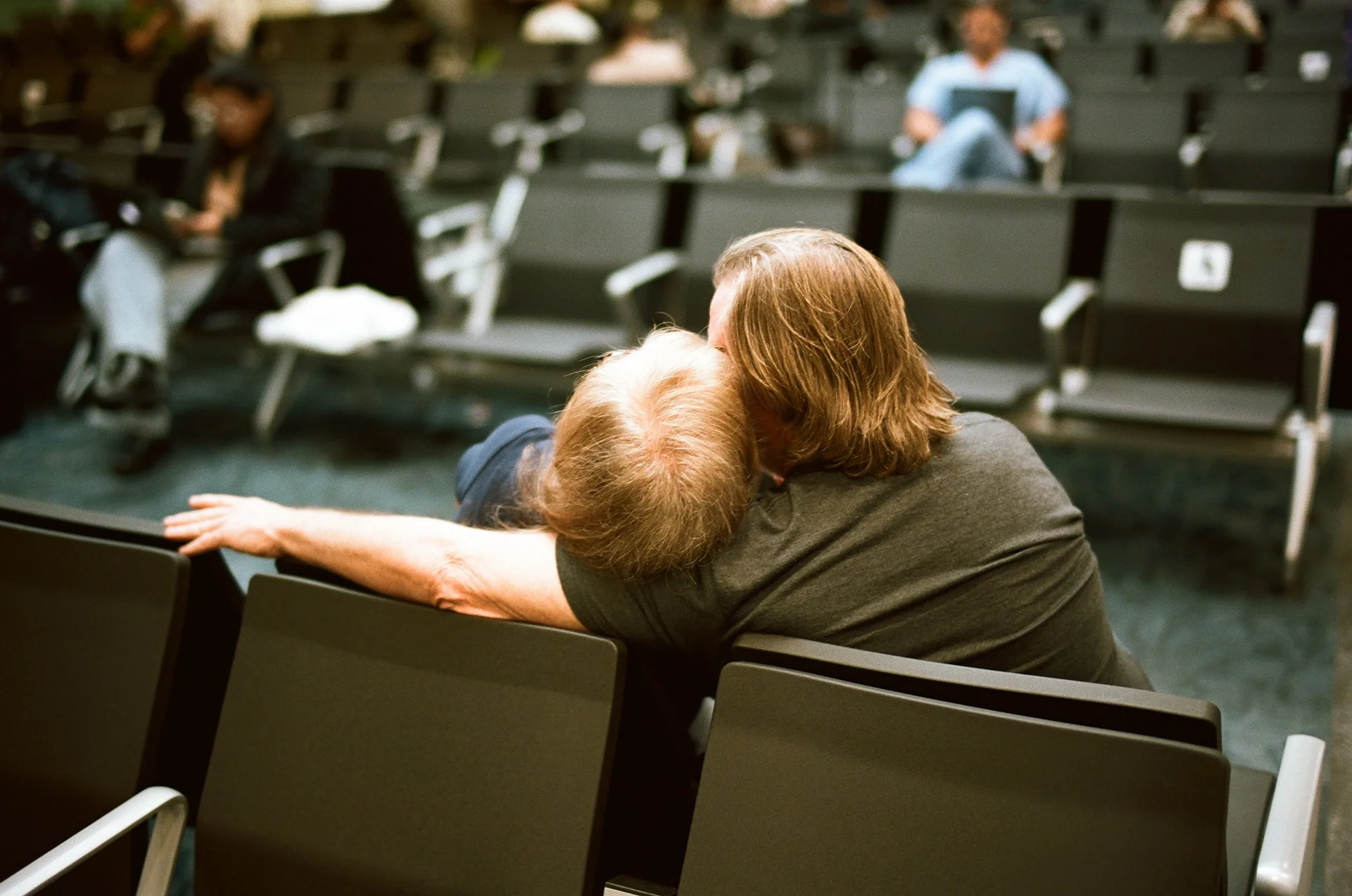 A person with shoulder-length brown hair is leaning over a desk, kissing a young child with light brown hair on the forehead in a conference or waiting room with rows of empty black chairs. Other people are seated in the background, some using laptop
