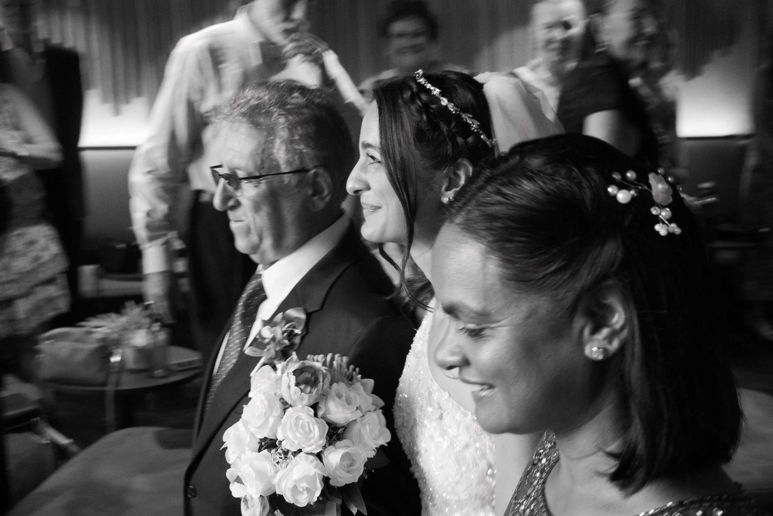 Black and white photo of a wedding ceremony showing a bride and groom sitting side by side, surrounded by guests. The bride is smiling, wearing a wedding dress with lace details, and has floral decorations in her hair. The groom is wearing glasses an