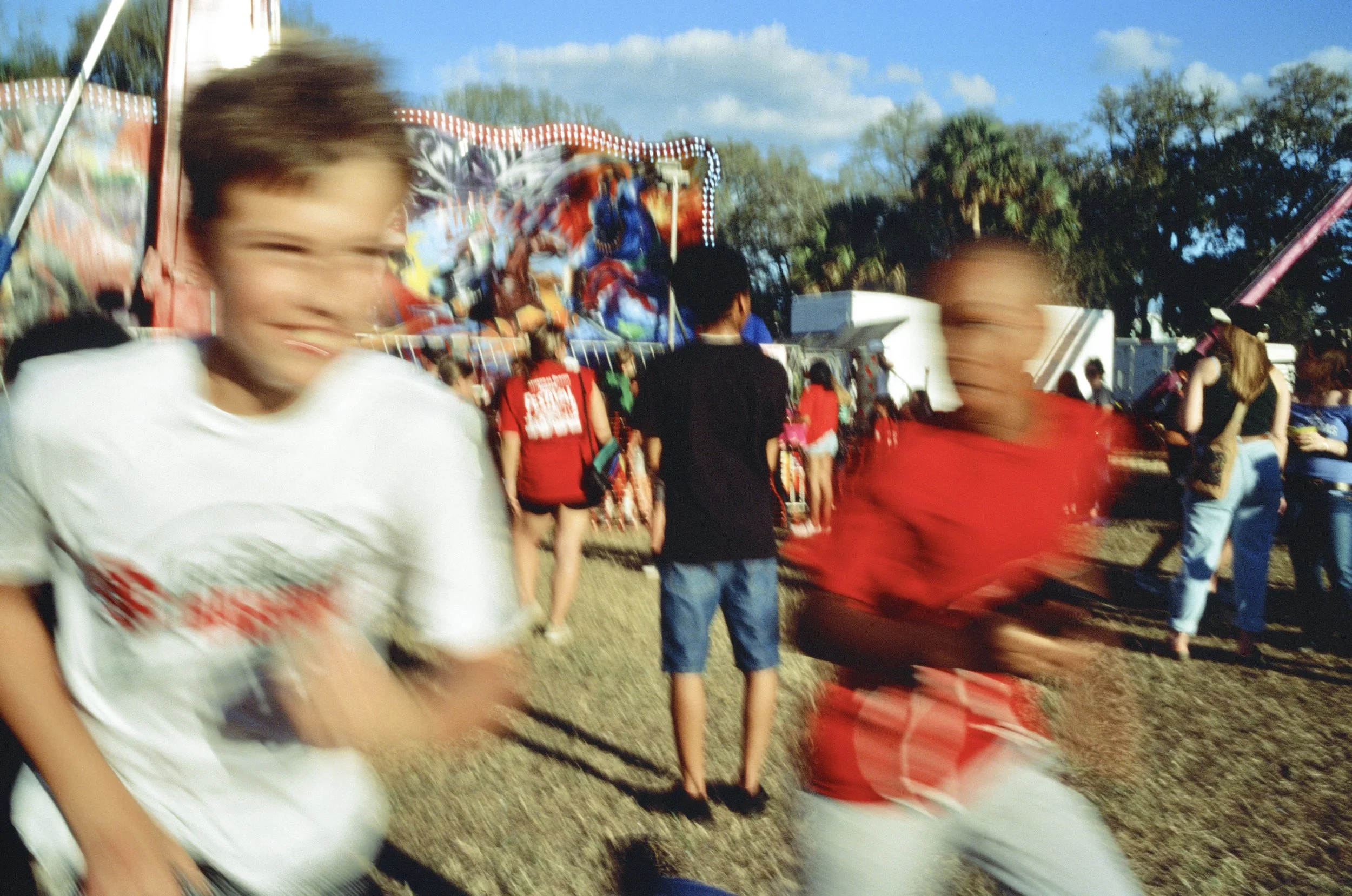 A crowded outdoor fairground with moving people, carnival rides, and a colorful painted mural in the background. The scene is lively with children and adults.