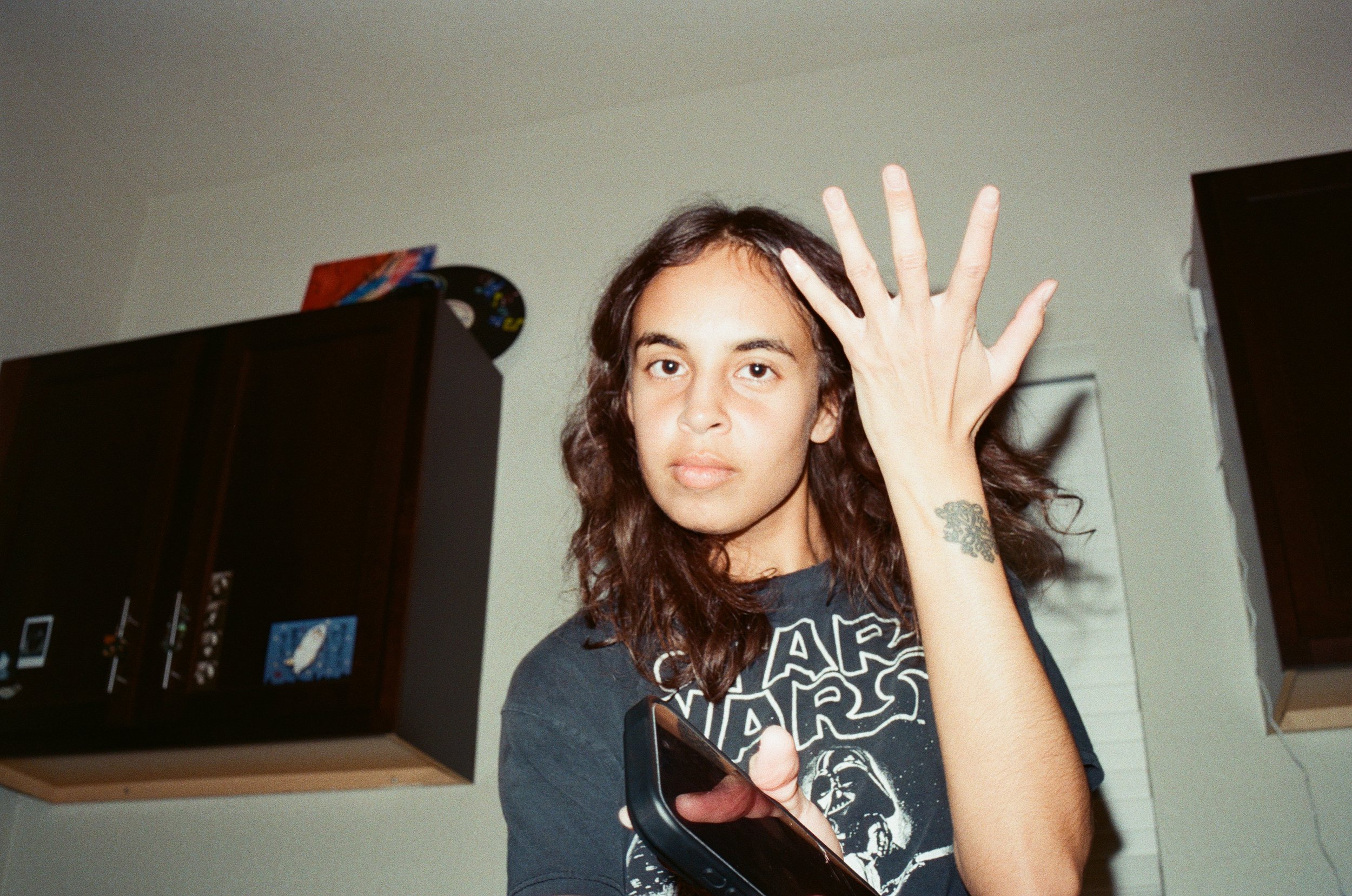 A young woman with long curly hair holding a phone in one hand and waving with the other, wearing a Star Wars t-shirt, standing in a room with dark furniture and light-colored walls.