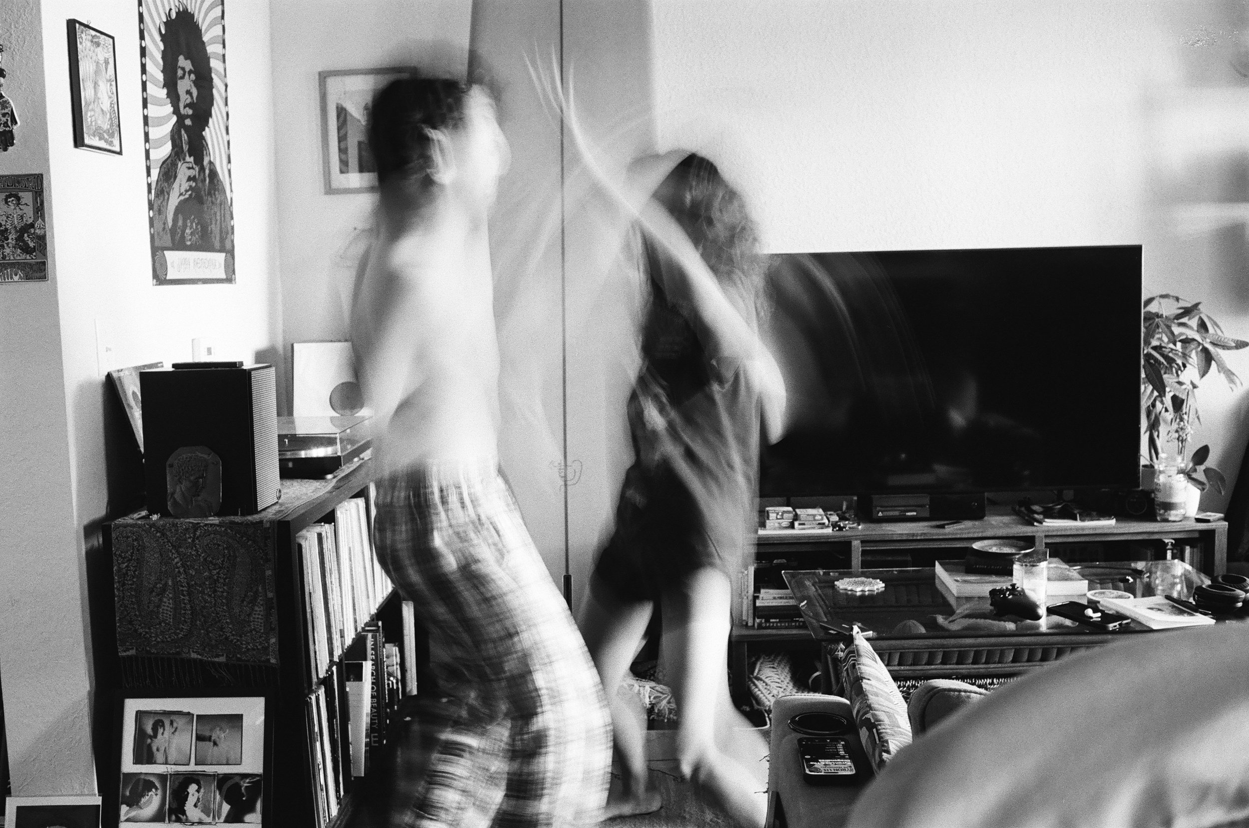 A black and white photo of two people dancing in a living room. The person on the left is wearing plaid pants and a sleeveless shirt, while the person on the right is dressed in dark clothes. The background features a television, a bookshelf filled w