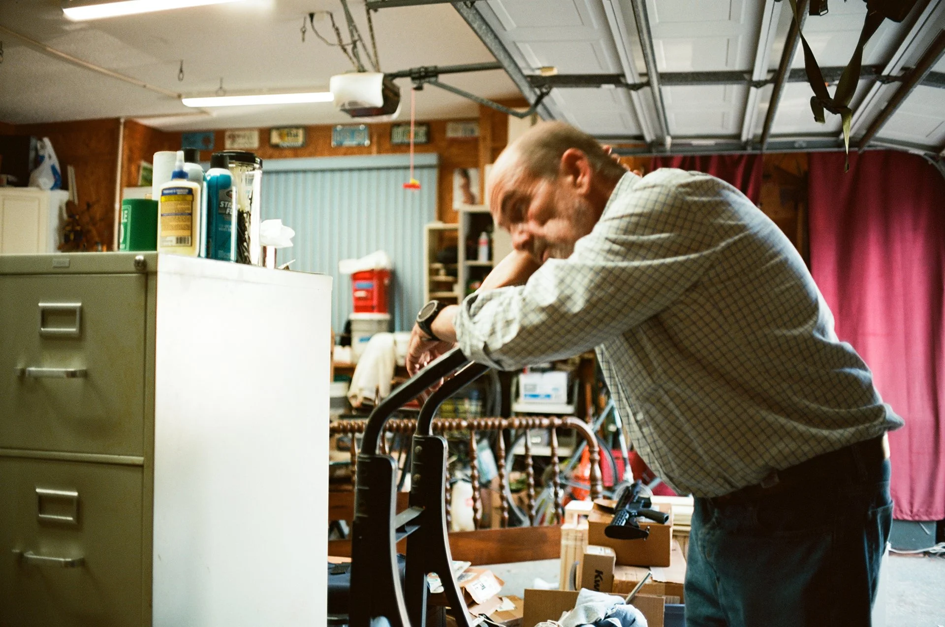A man leaning on a walker in a cluttered garage, surrounded by shelves and various items, with red curtains in the background.