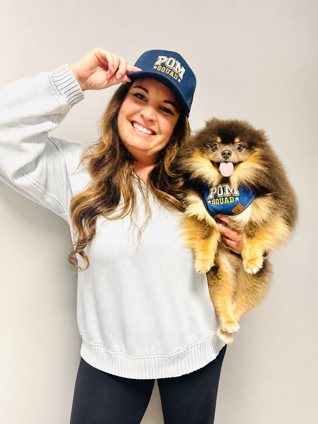 A smiling woman wearing a "POM Squad" baseball cap holding a fluffy Pomeranian dog with a "POM Squad" bandana.