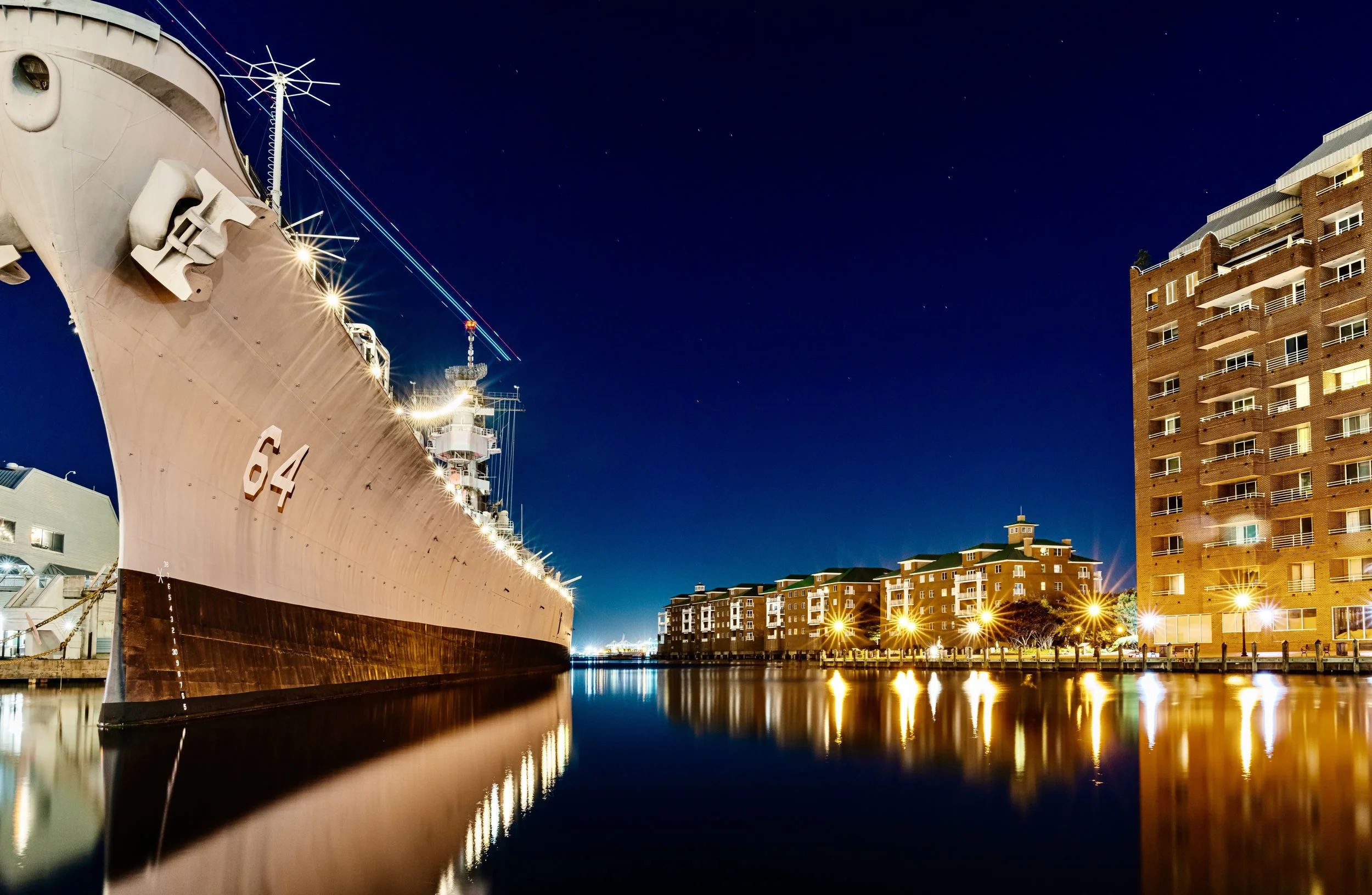 Night view of a large ship docked beside modern apartment buildings, with reflections in the water and a clear starry sky above.