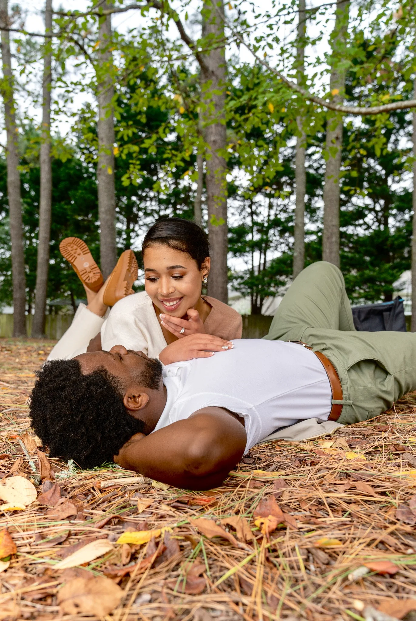 A woman and a man lying on the ground in a wooded area, with the woman smiling and looking at the man, who appears to be resting with his eyes closed.
