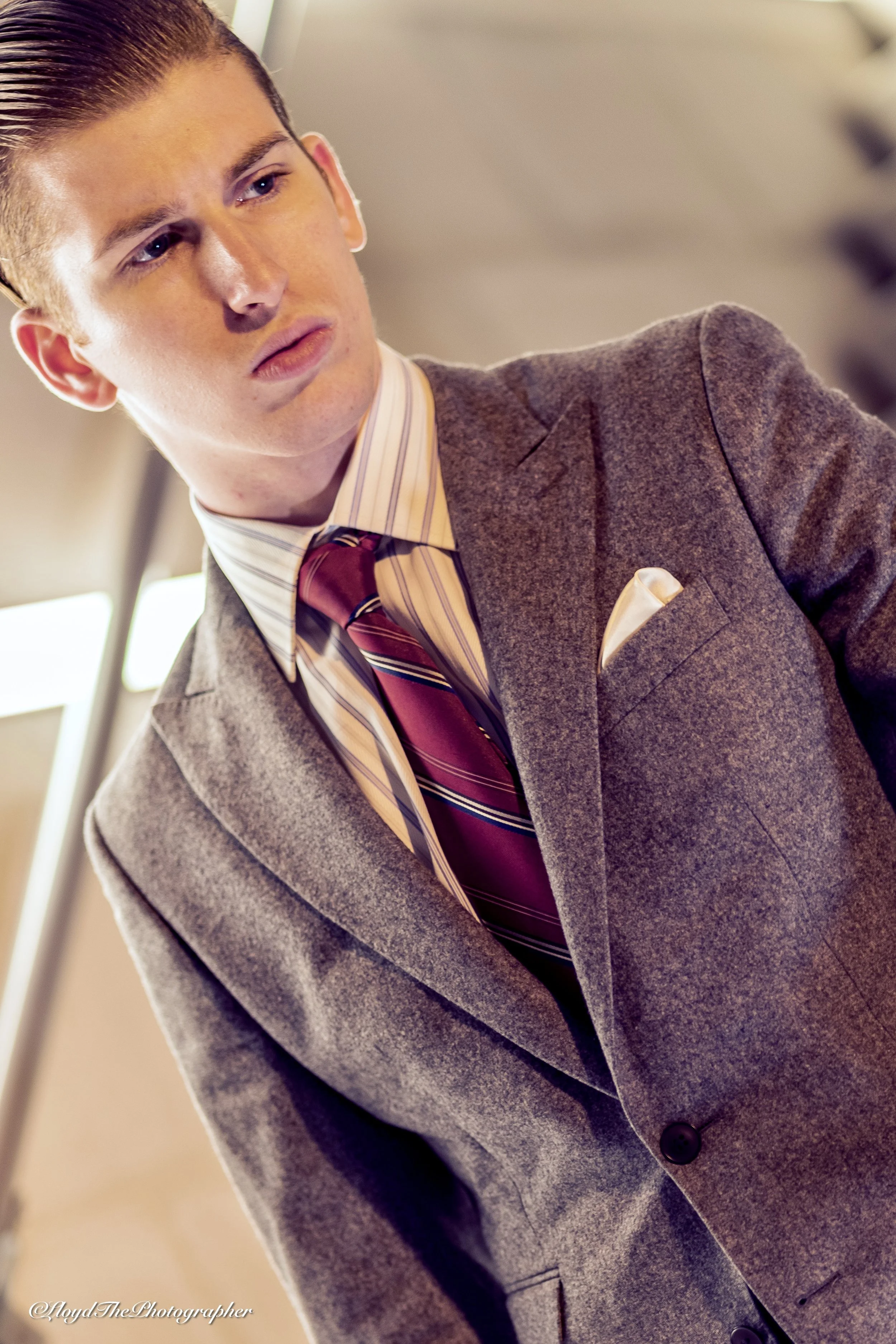 A young man with slicked-back hair wearing a gray suit, striped shirt, red and navy tie, and pocket square, standing indoors with a blurred background.