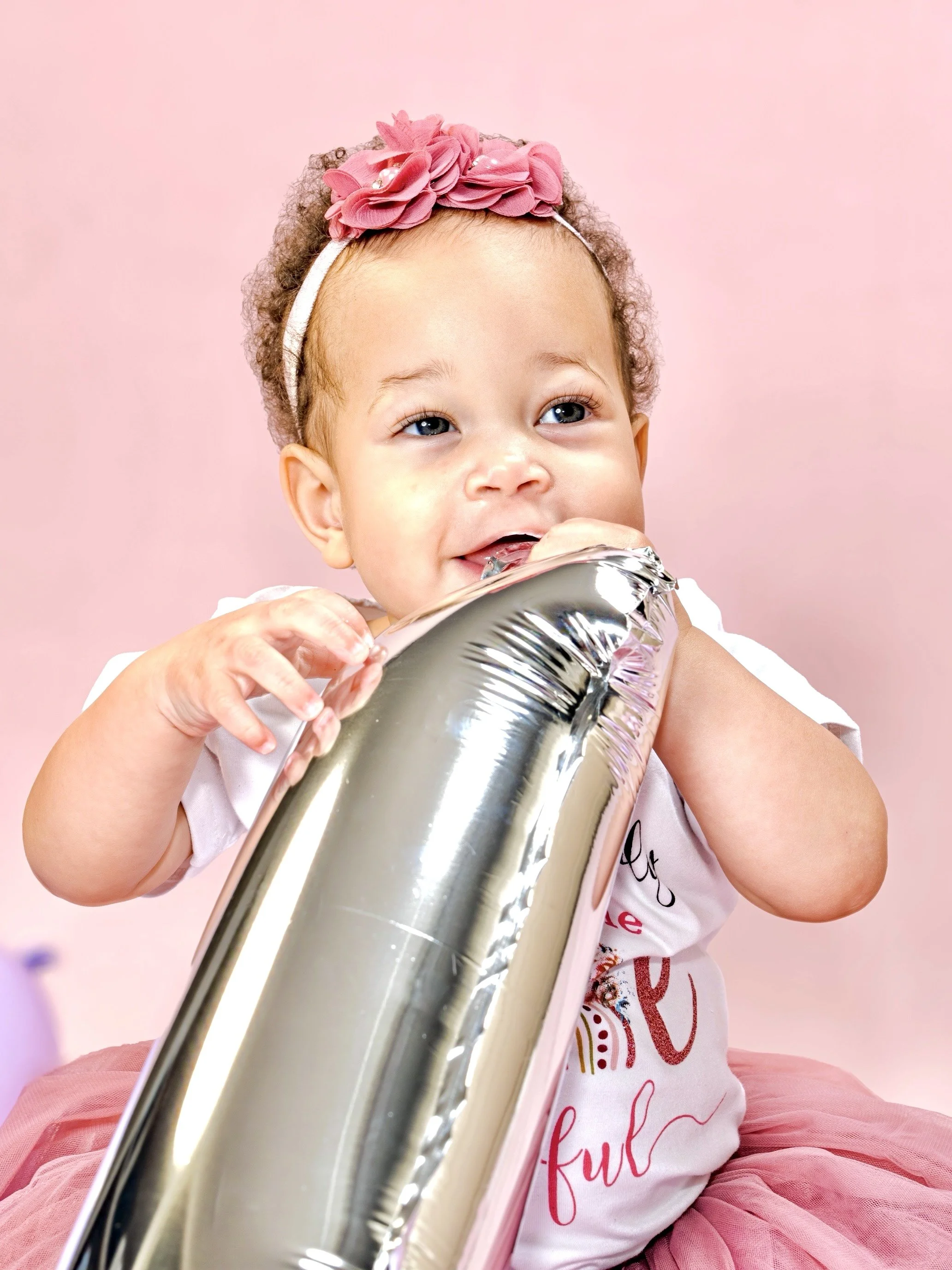 A young girl with curly hair wearing a pink headband with fabric flowers, biting a shiny metallic balloon, against a pink background.