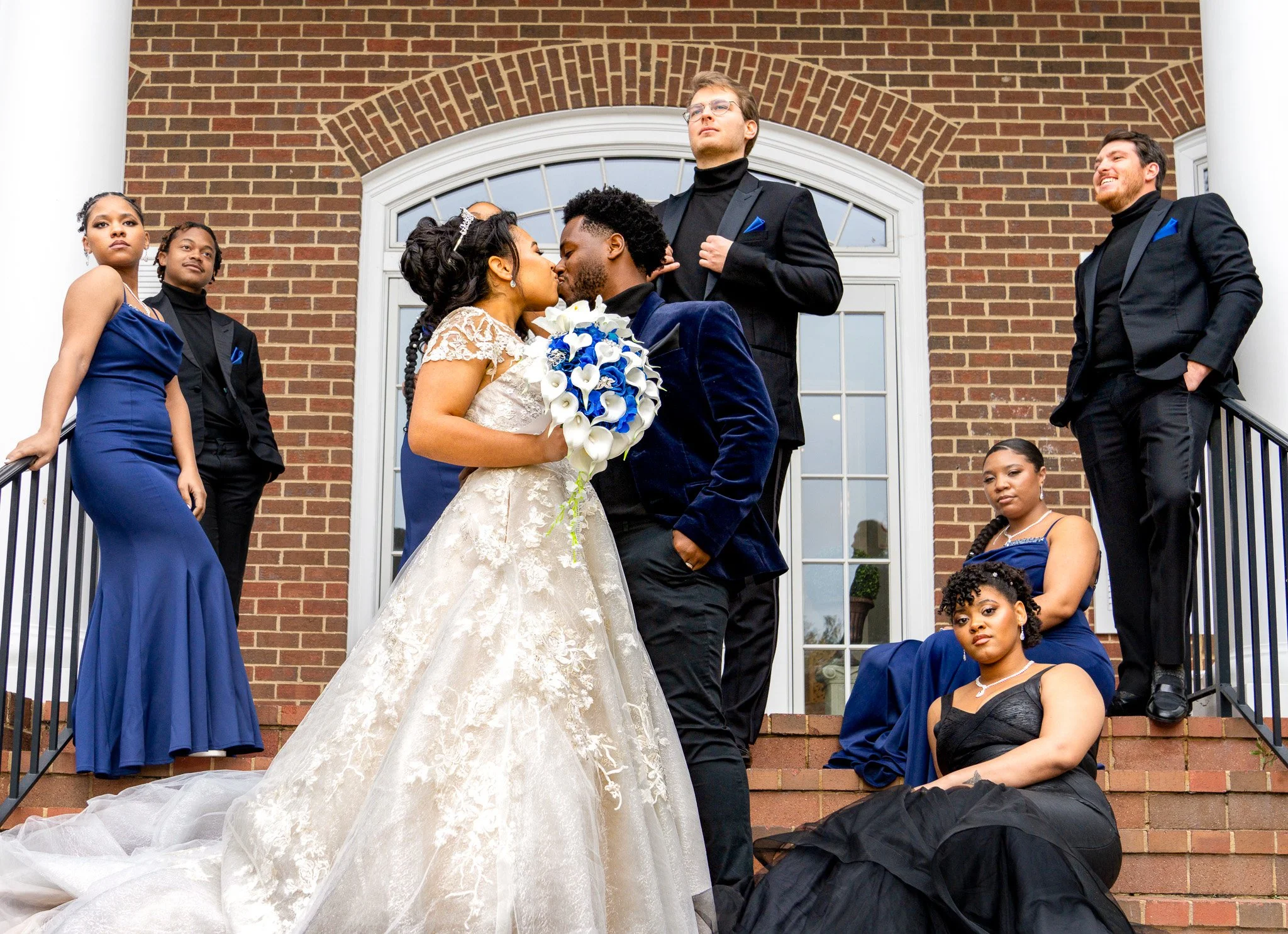 A diverse group of six people, including a bride and groom, standing on the steps of a brick building with large white columns, during a wedding celebration. The bride in a white gown and the groom in a dark suit are kissing, while the others pose ar