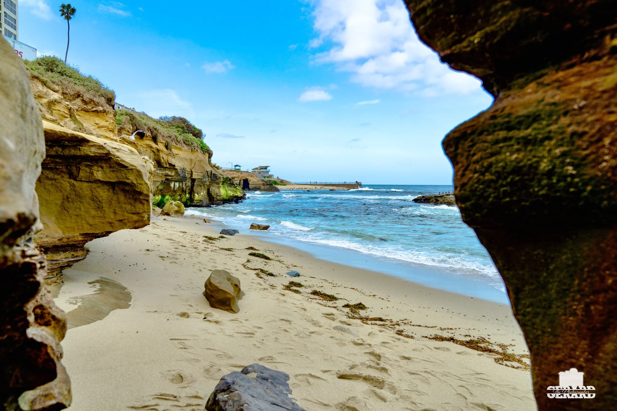 View of a sandy beach through rock formations with cliffs, ocean waves, and blue sky with clouds.