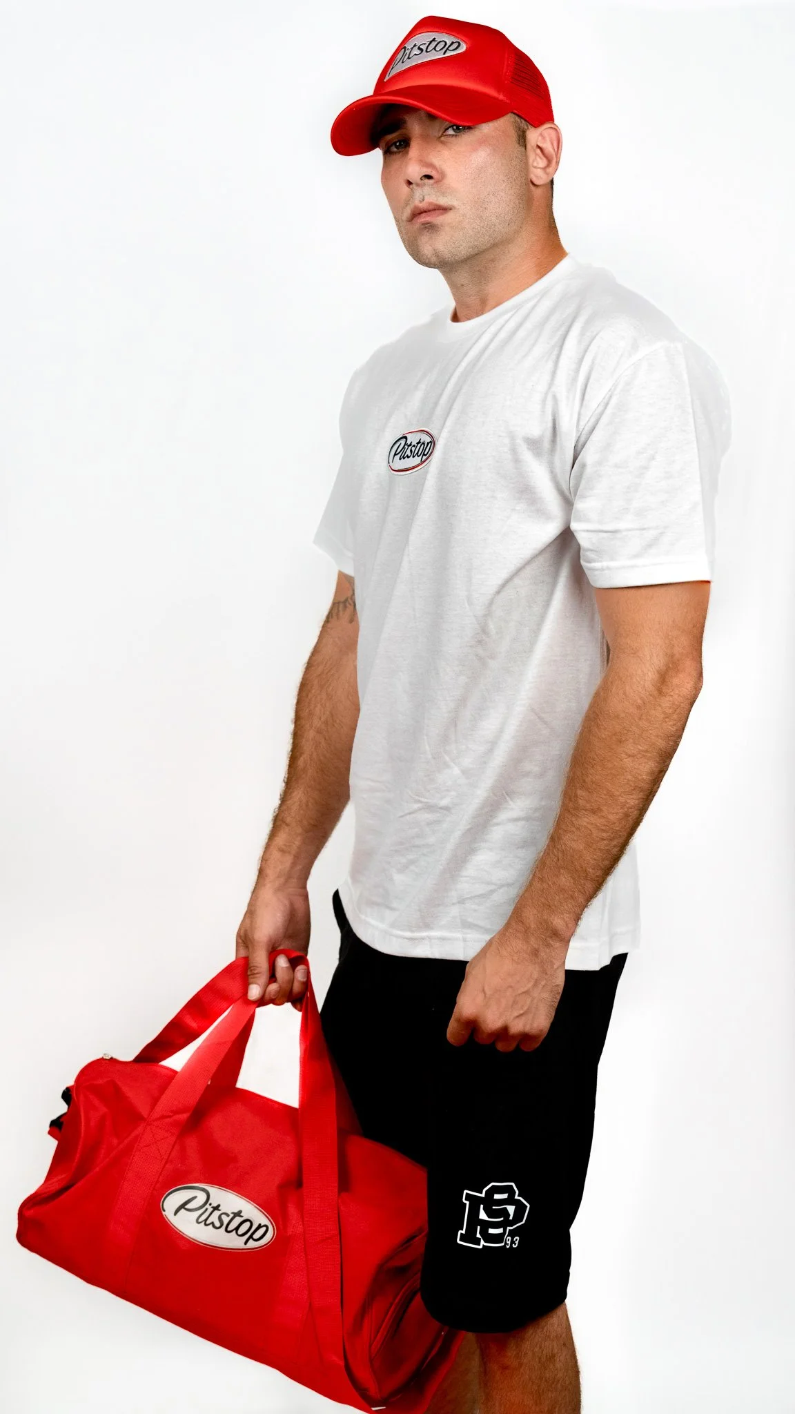 A man wearing a red cap with the word 'Pitstop' and a white T-shirt, holding a red bag with 'Pitstop' logo, standing against a plain white background.