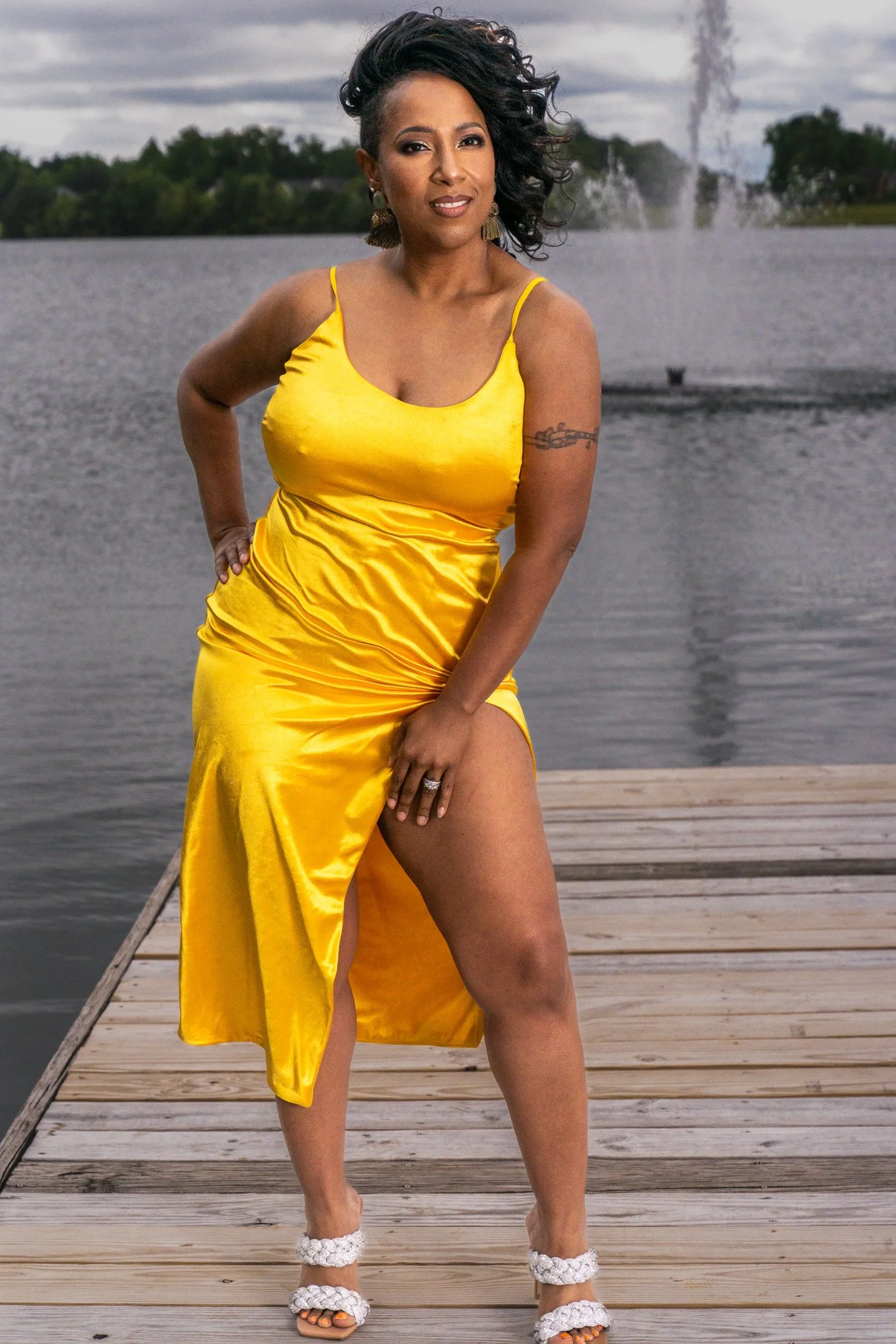 A woman with dark, curly hair styled in a voluminous side-swept manner, wearing a bright yellow satin dress with thin straps, standing on a wooden dock by a lake. She has large earrings, a tattoo on her upper arm, and is posing with one hand on her h