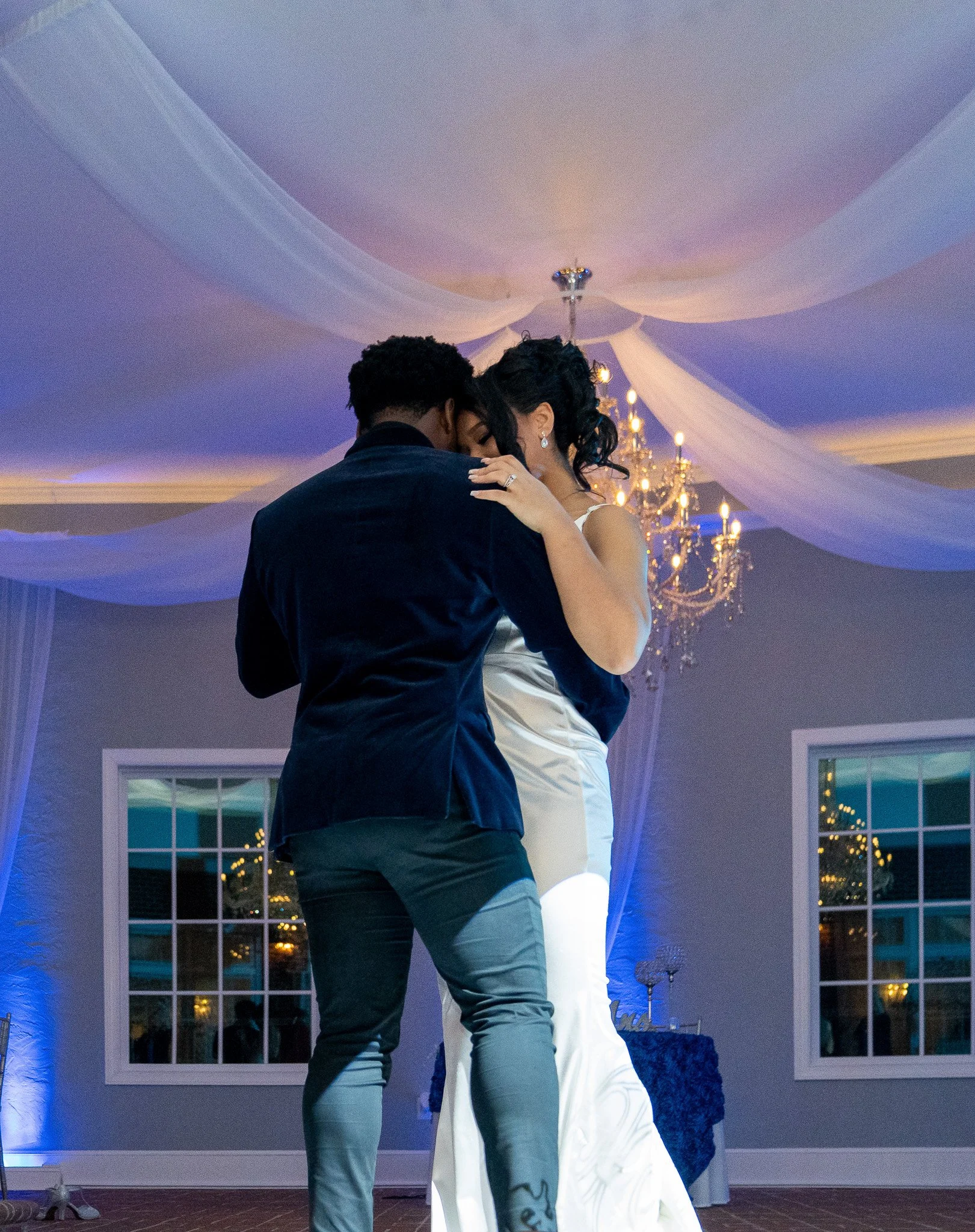 A bride and groom sharing a dance at their wedding reception, with the bride wearing a white gown and the groom in a dark suit, under decorated ceiling and chandelier lights.