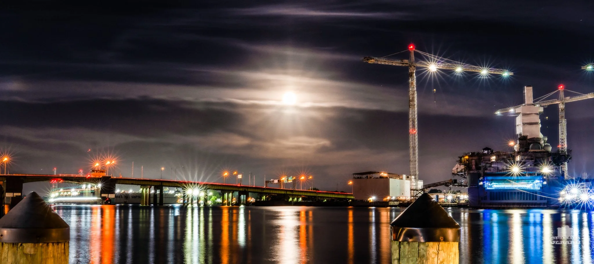 Nighttime scene of a construction site by the water with two large cranes, illuminated by bright lights, with a full moon and cloudy sky in the background, and reflections on the water surface.