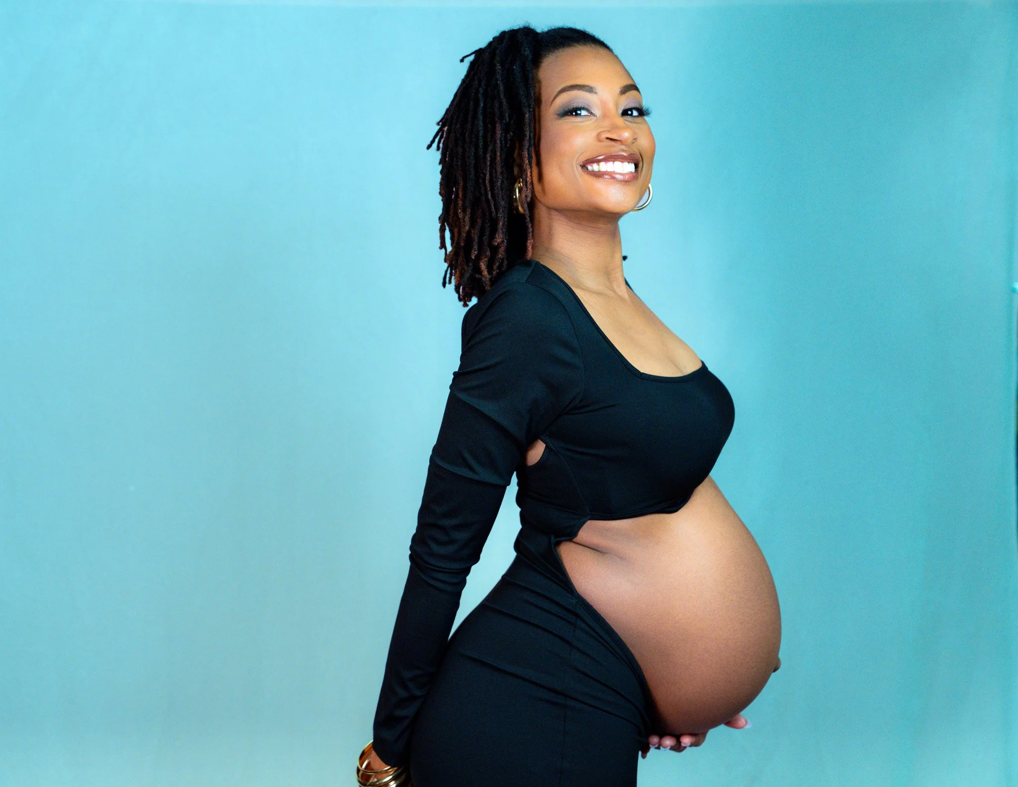 A pregnant woman with dreadlocks and gold hoop earrings smiling against a light blue background.