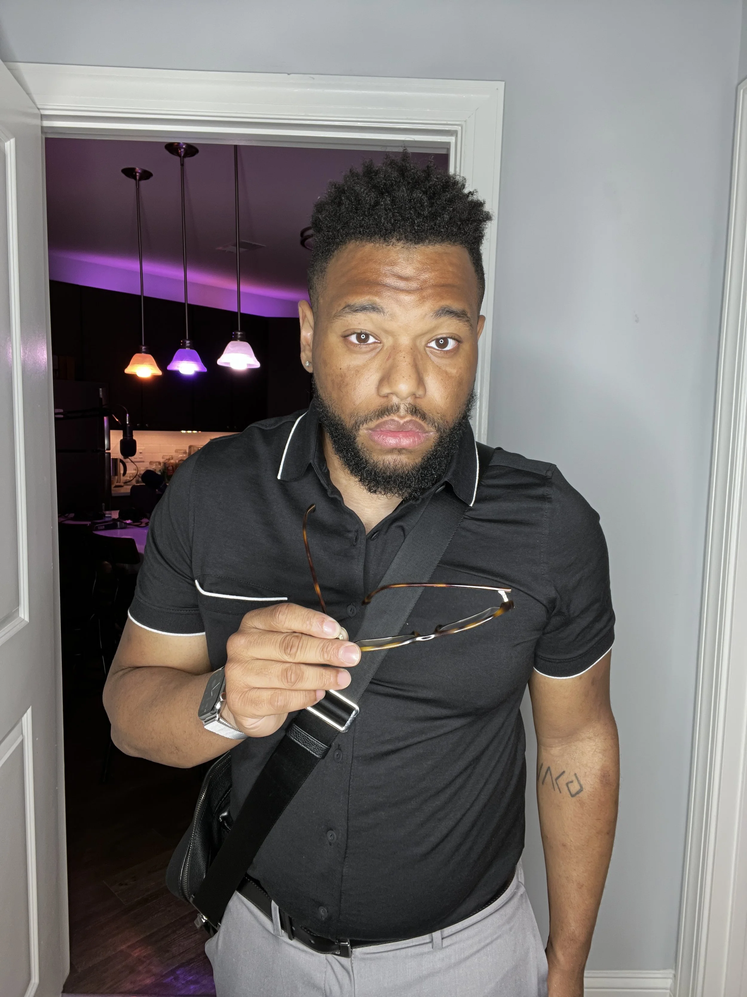 A young man with a beard and short curly hair standing in a doorway holding glasses, wearing a black polo shirt, gray pants, and a smartwatch on his left wrist, with a black crossbody bag.