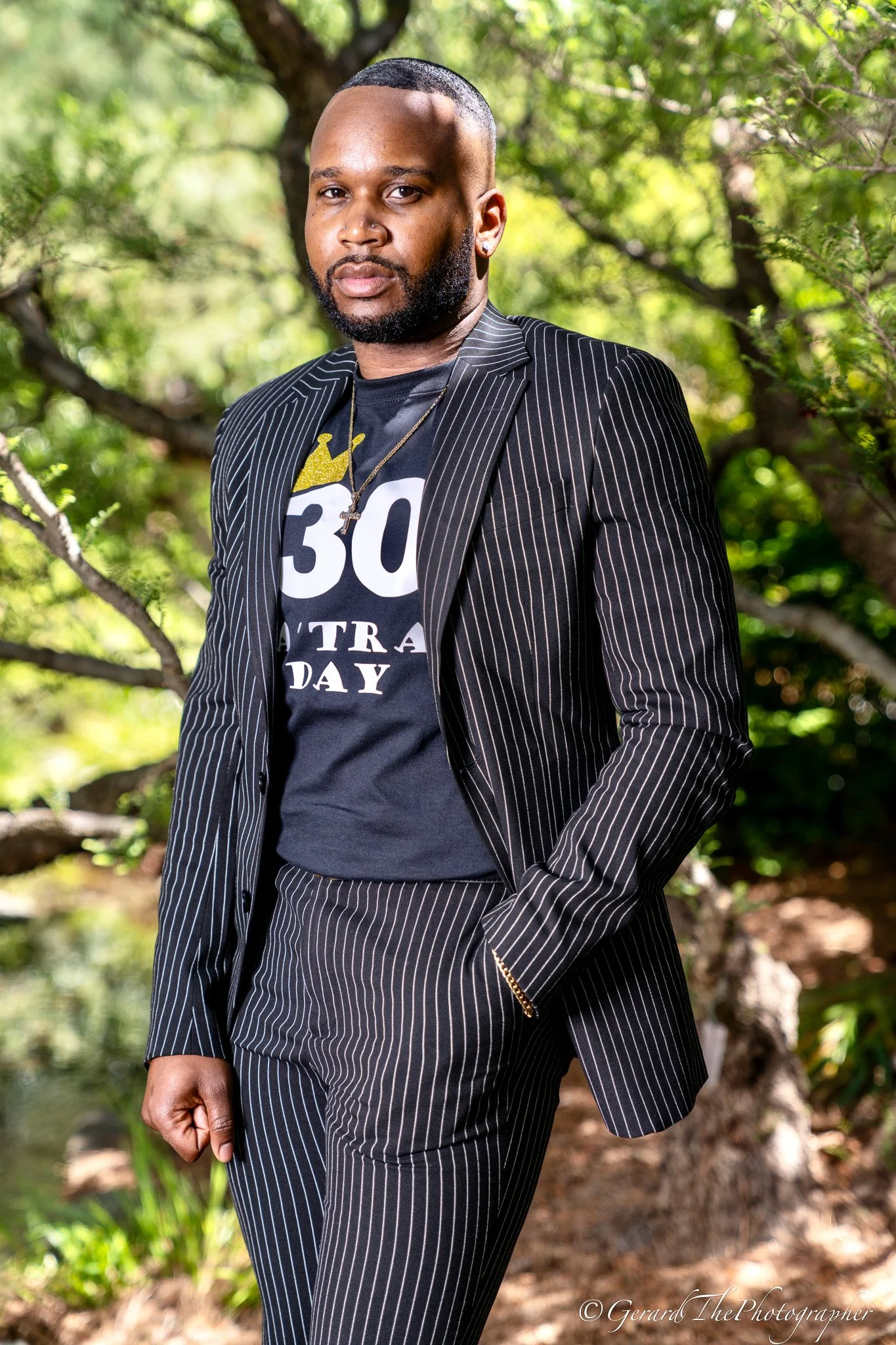 A man with a beard and pierced ears standing outdoors near a tree, wearing a black pinstripe suit jacket over a black graphic t-shirt with the number 30 and a crown. The background features trees and greenery.